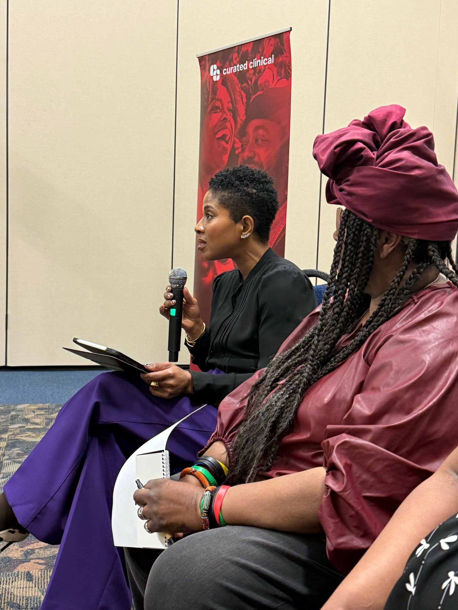 Person with braided hair and colorful bracelets sitting with a notebook, holding a pen, and a person in a black shirt holding a microphone, seated against a beige wall with a red poster.