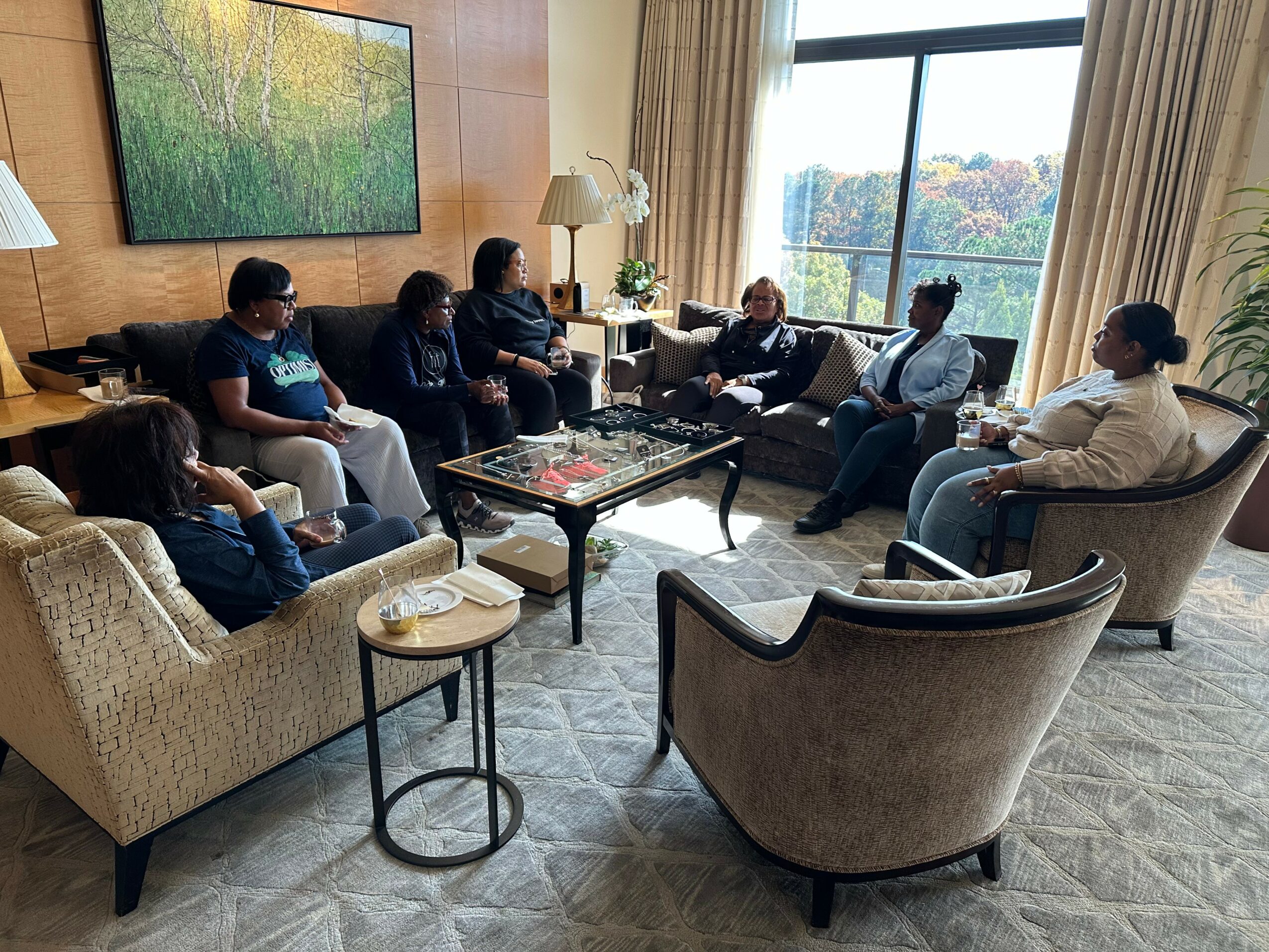 Group of seven people sitting in a living room with large window, some wearing masks, engaged in conversation.