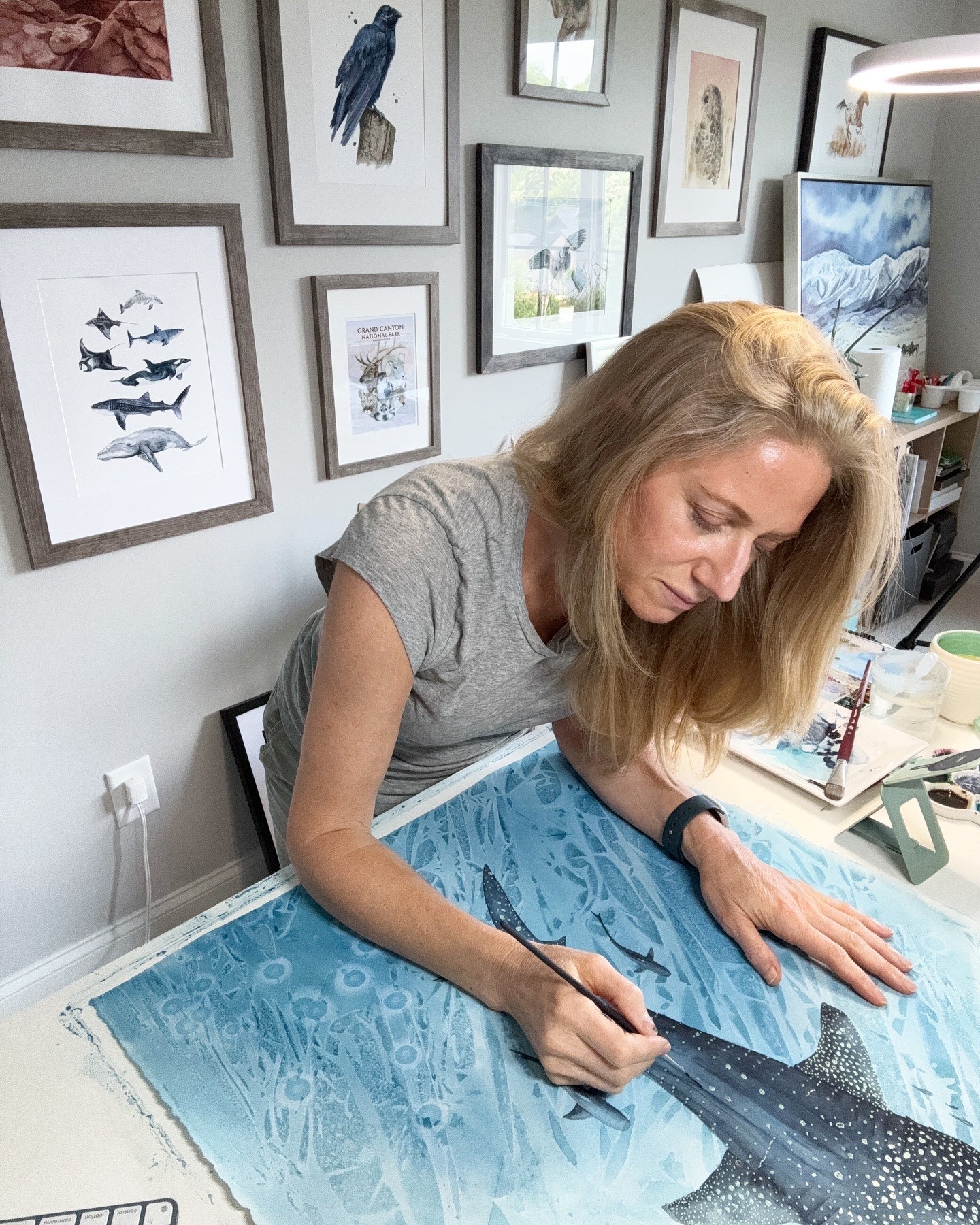 Woman painting a large blue and black whale on a table, surrounded by framed artwork on the wall.