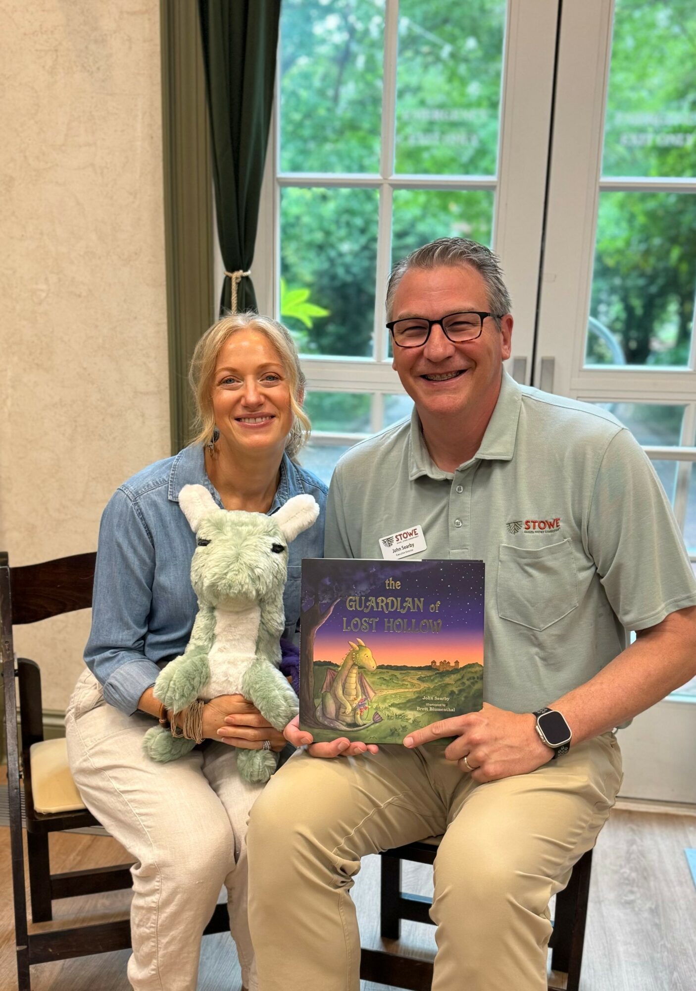 Two people sitting indoors near a window, holding a children's book and a stuffed animal, smiling at the camera.
