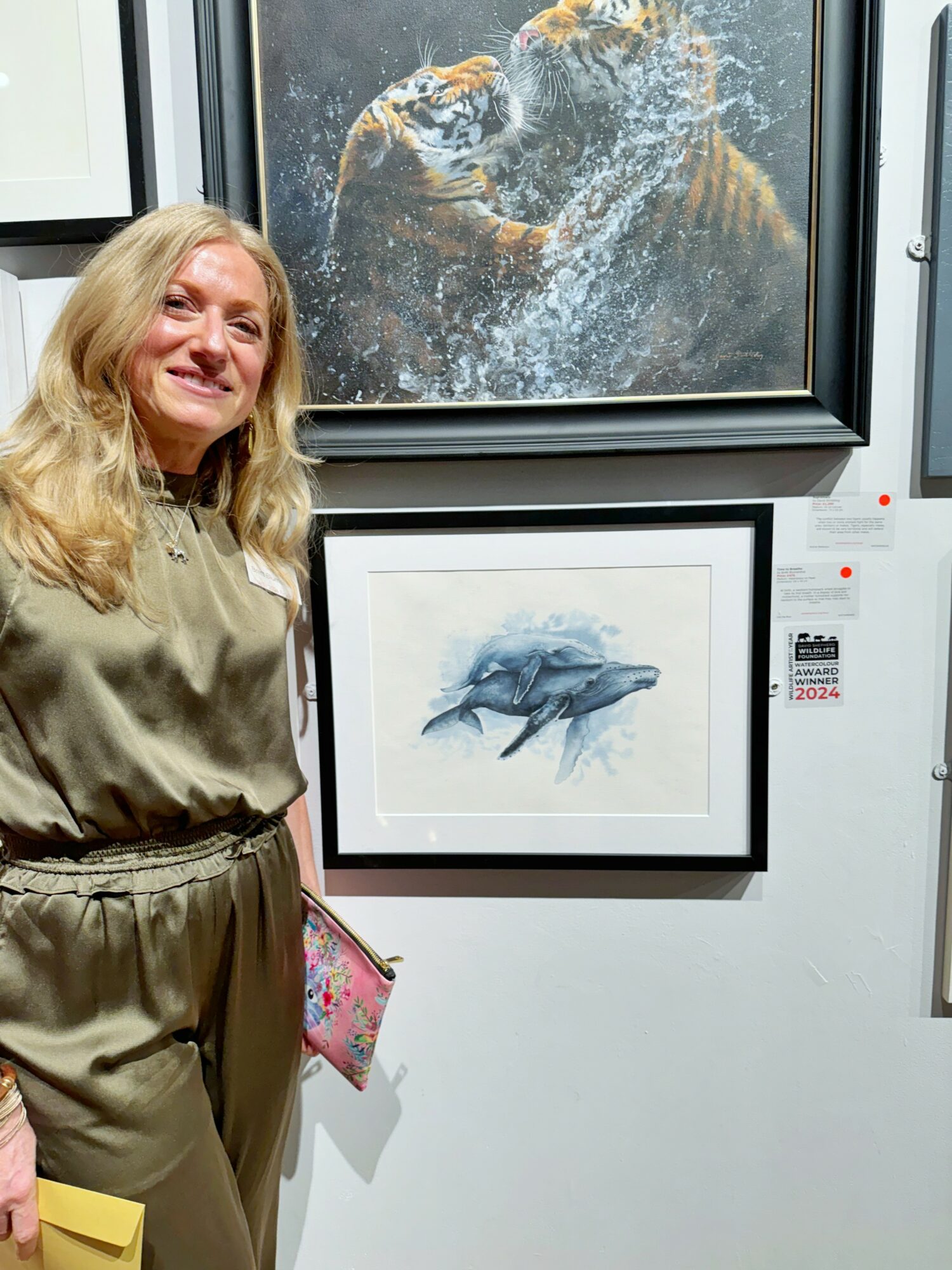 Woman with long blonde hair standing next to framed artwork of two dolphins jumping out of water, in an exhibition setting.
