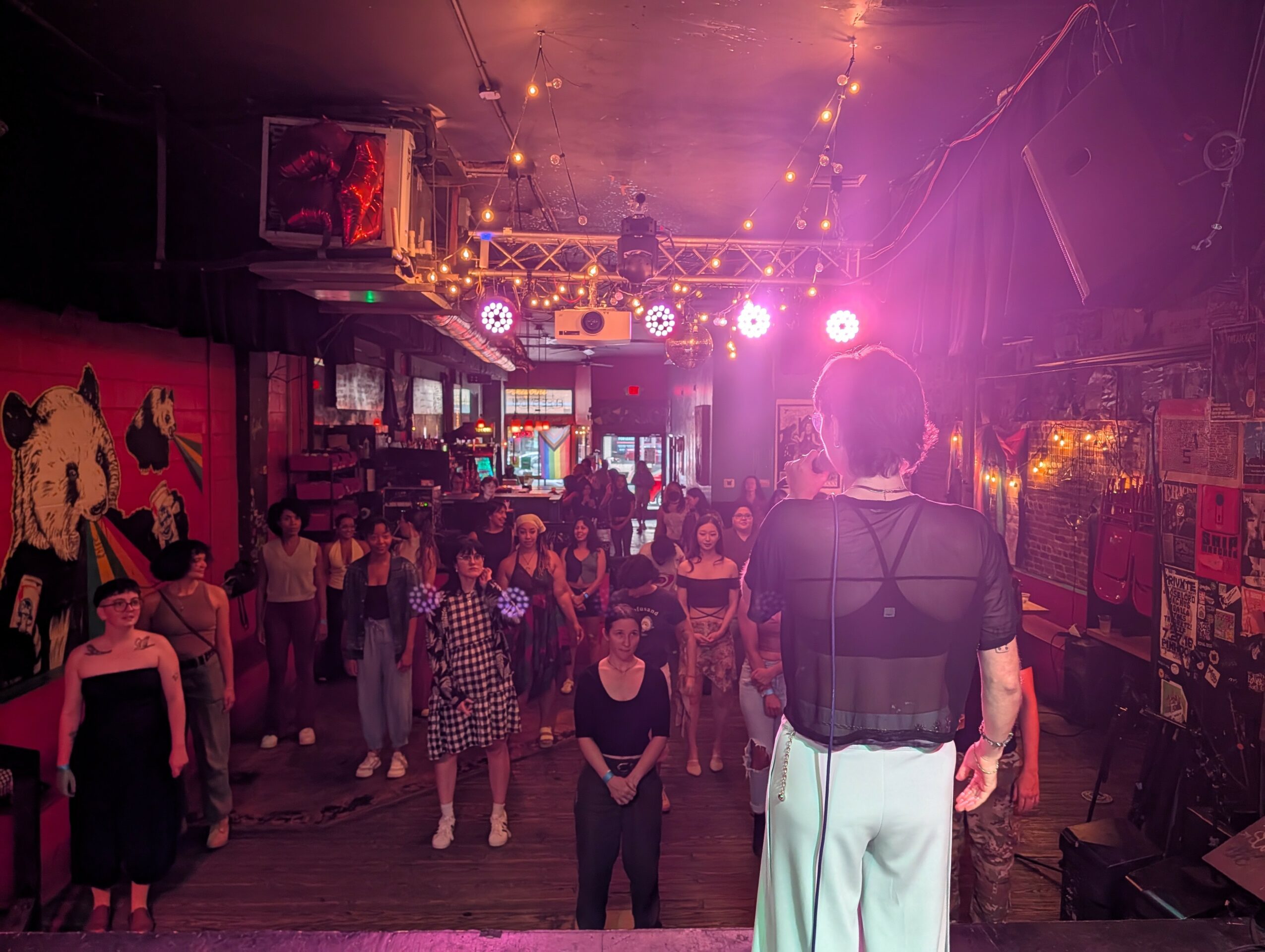 Performer on stage facing audience in a dimly lit venue with colorful lights and posters on walls.