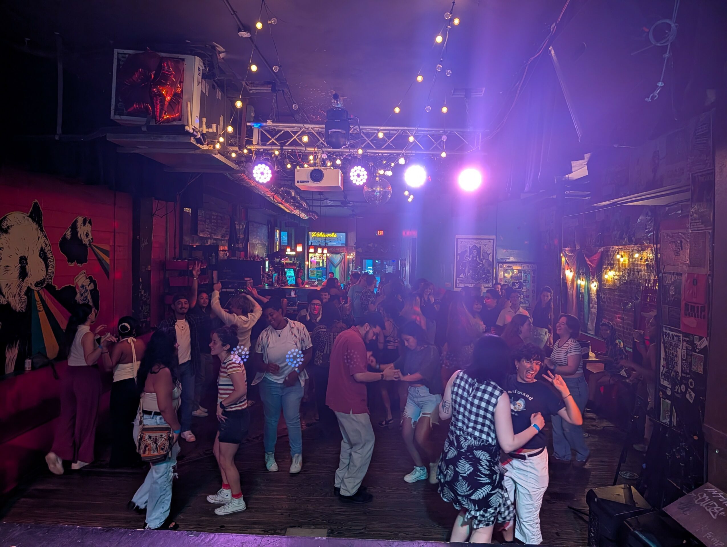 People dancing and socializing in a dimly lit nightclub with colorful lights and artwork on walls.