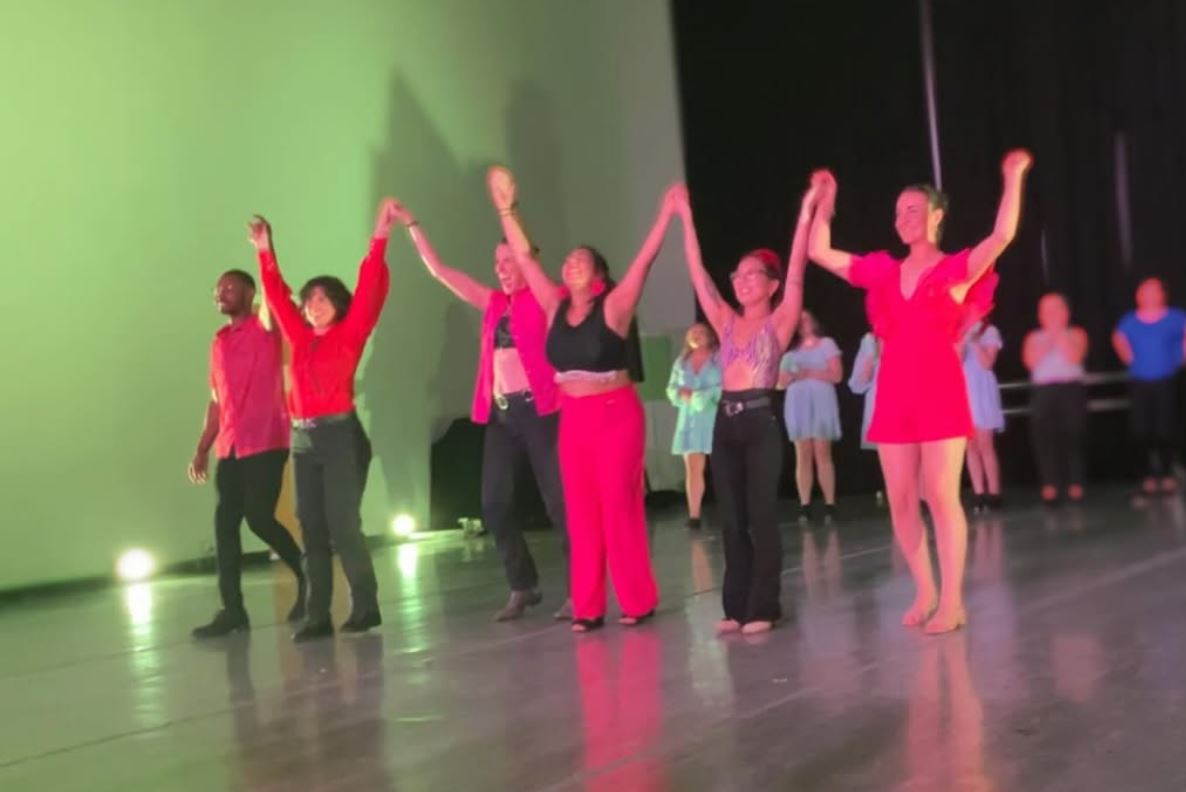 Group of dancers on stage holding hands, raising arms, with a green and black background, some in pink and red costumes.