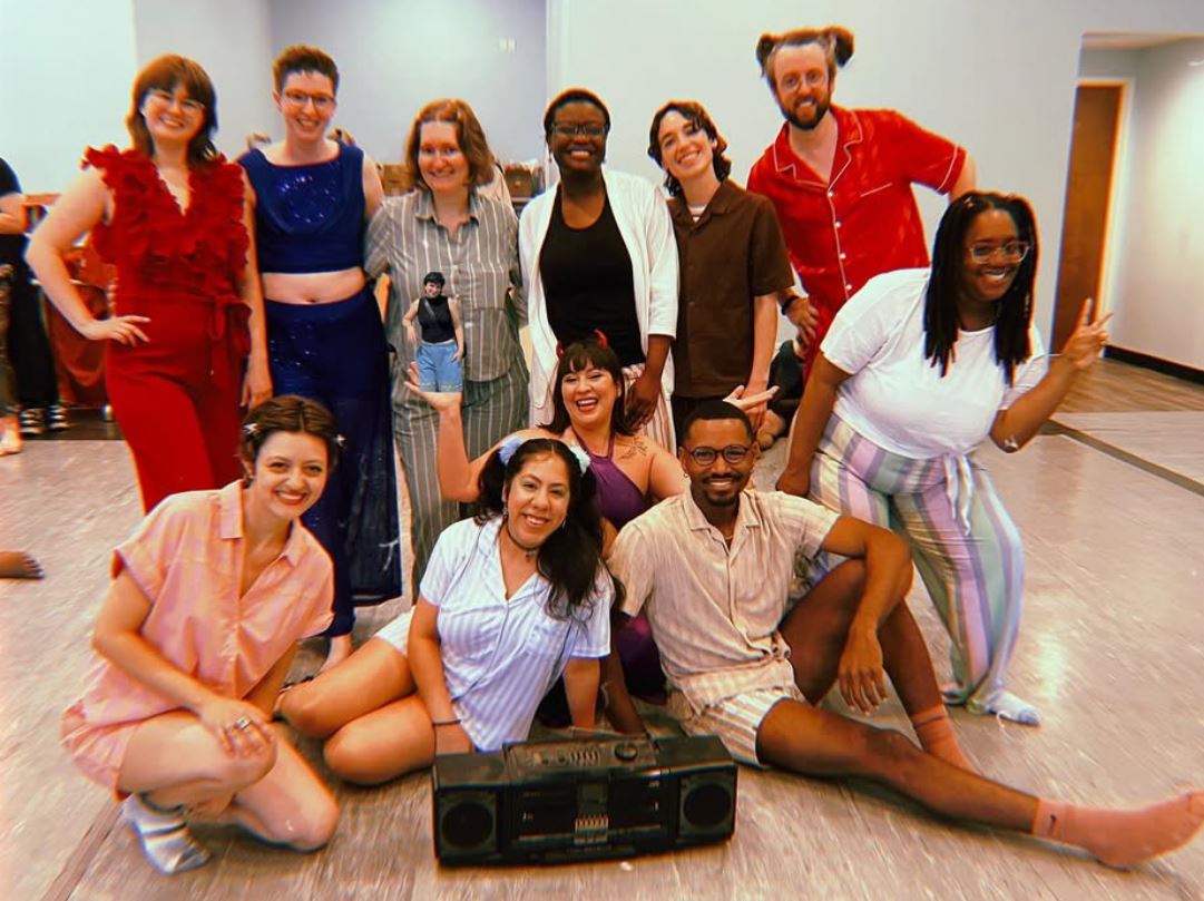 Group of ten people smiling and posing indoors, some sitting and some standing, with a boombox on the floor.