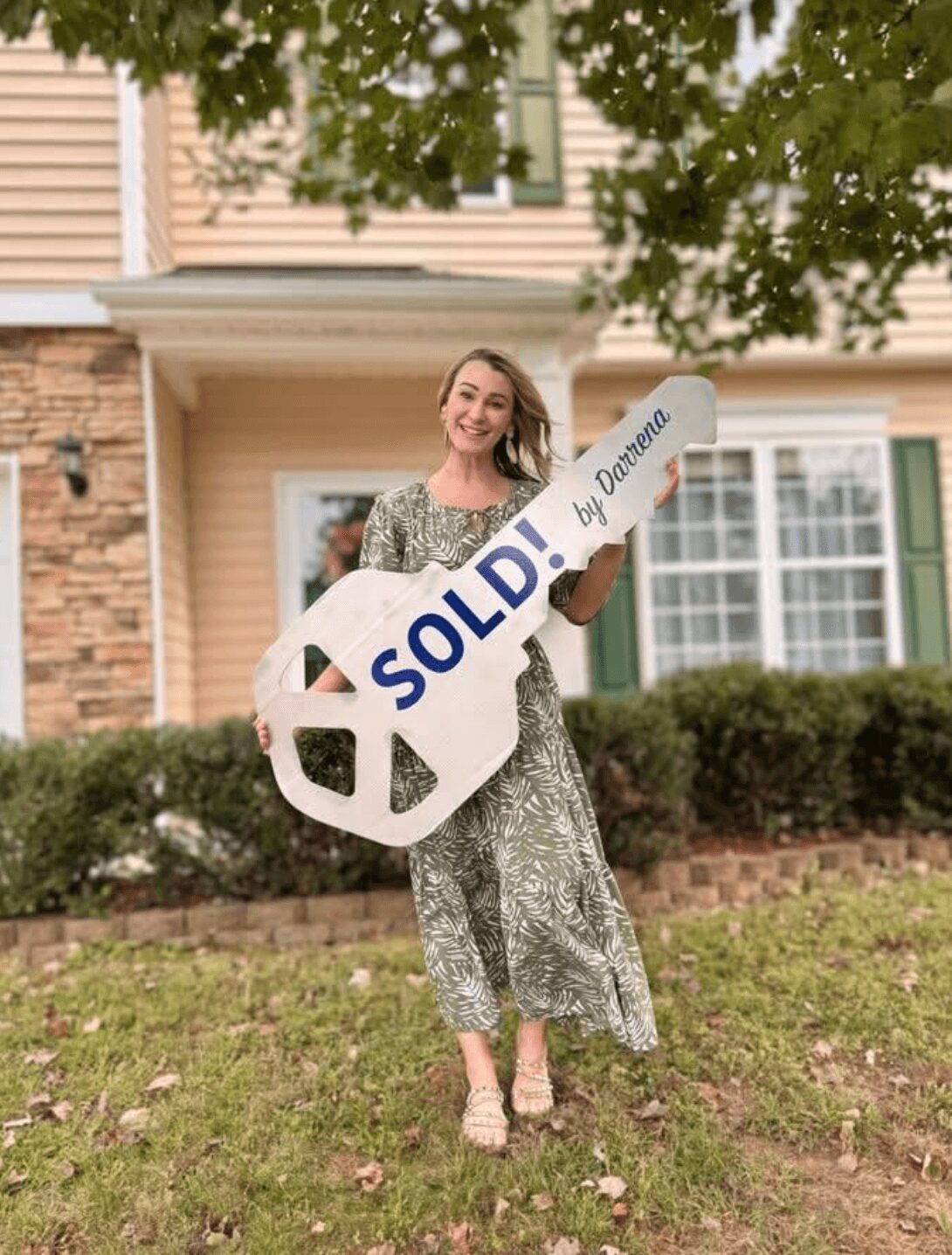 Woman holding a large key-shaped sign that says 'SOLD! by Domina' in front of a house with a lawn and trees.