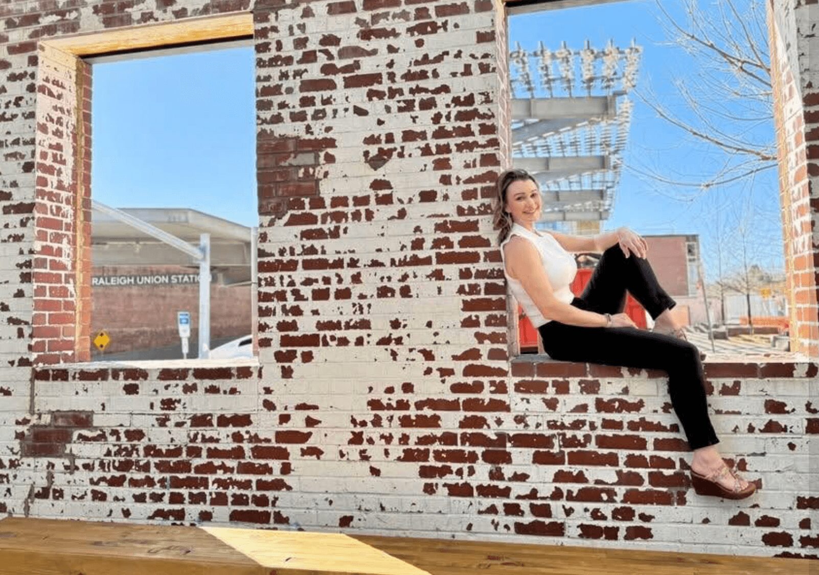 Woman sitting on a brick wall with windows behind her, outdoors, with a clear sky and building in background.