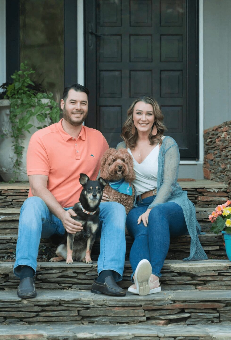 A smiling couple sitting on stone steps with two dogs in front of a house door, one large and one small, during daytime.