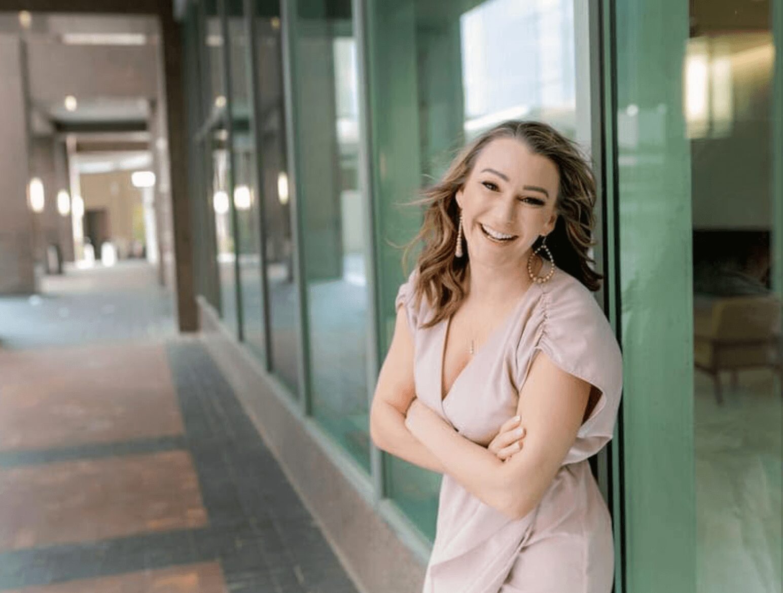 Smiling woman with wavy hair leaning against a glass wall in an indoor corridor.
