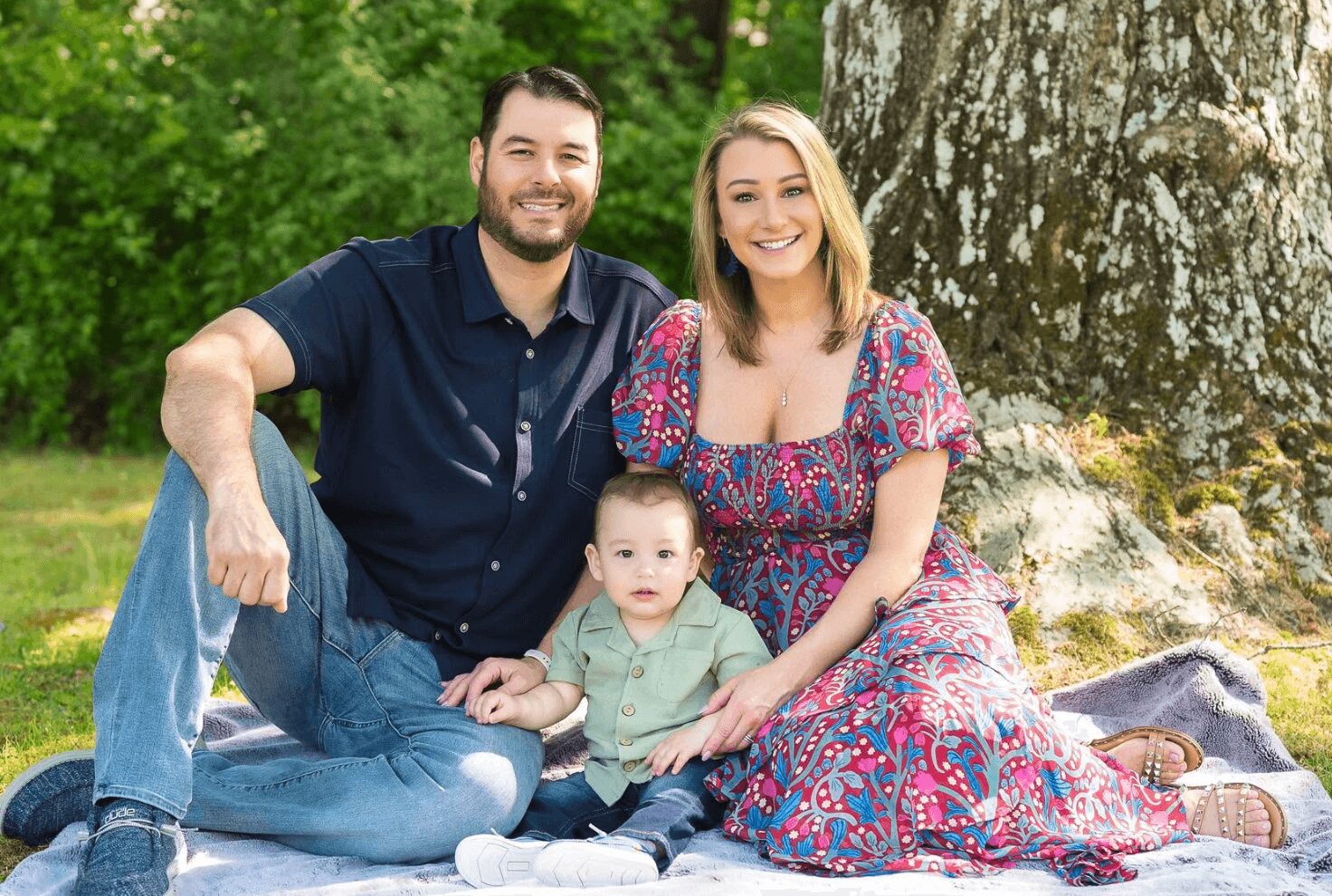 Family of three sitting outdoors on a blanket, smiling, with trees in the background.