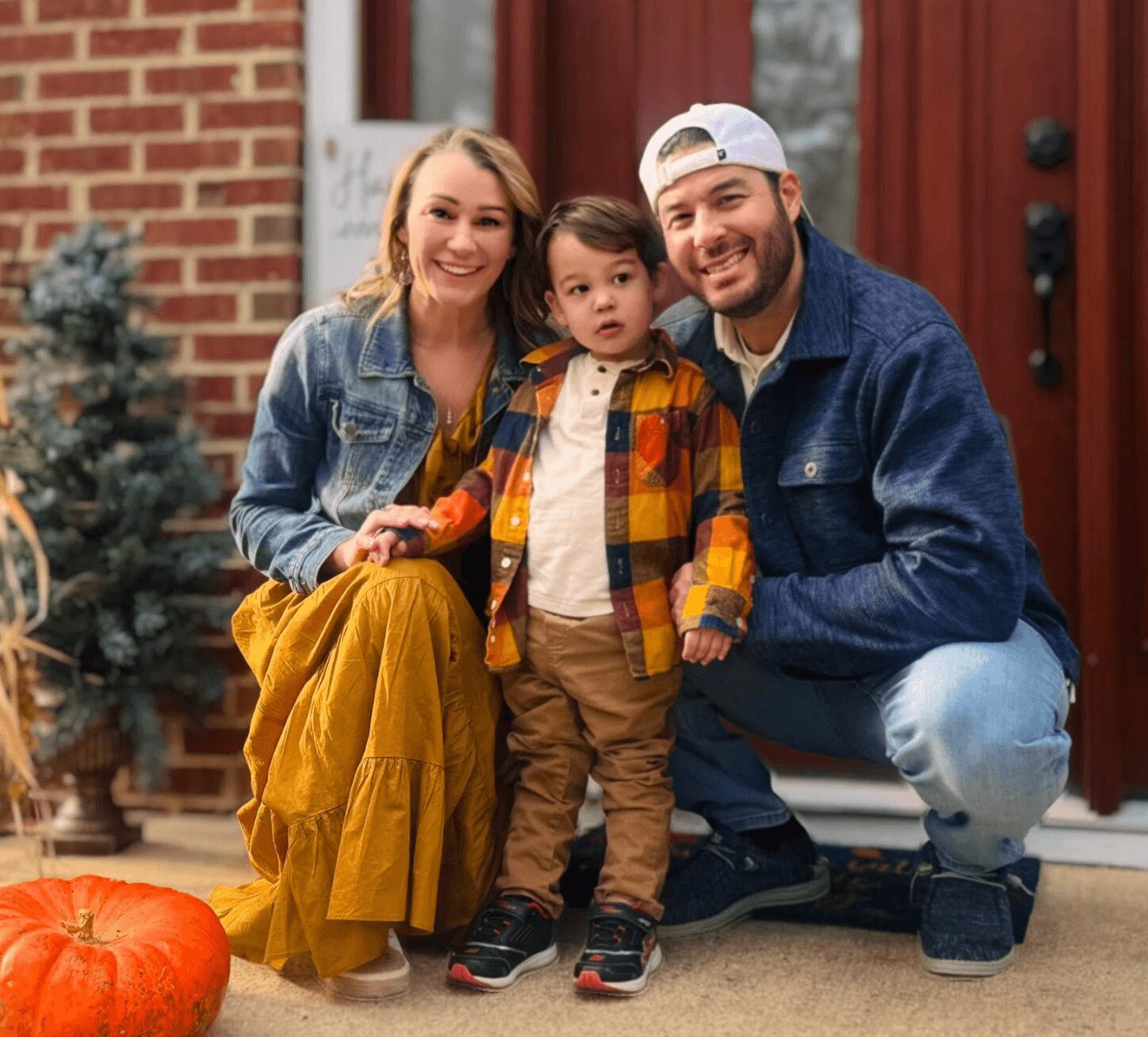 Family of three smiling outside house, child in colorful jacket, woman in yellow skirt, man in blue jacket, pumpkin nearby.