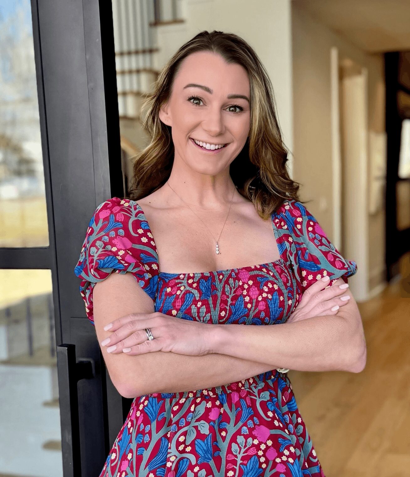 Smiling woman with arms crossed, wearing a colorful patterned dress, standing indoors near a glass door.