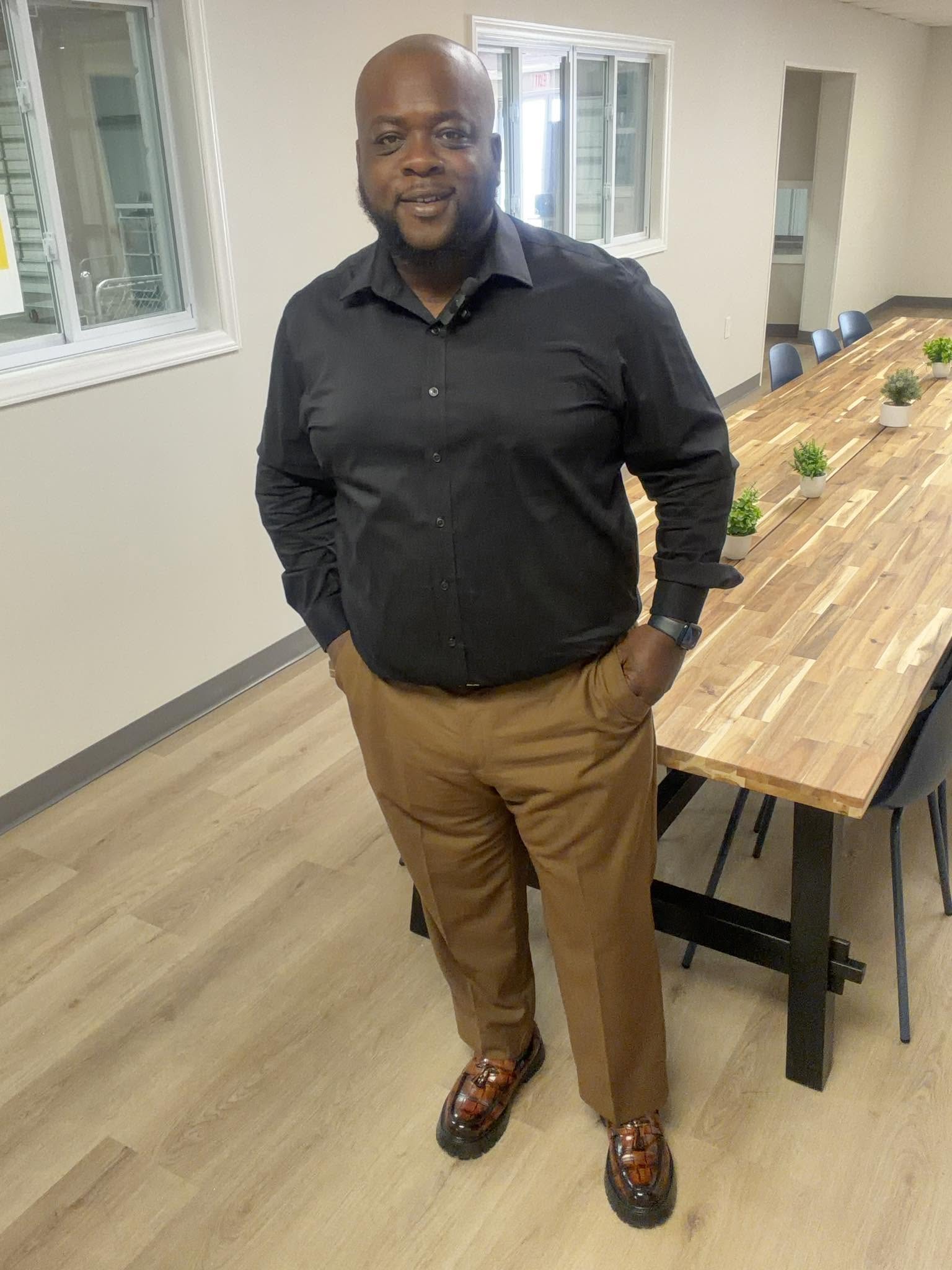 Man standing in a room with a long table, windows, and wooden flooring, smiling and looking at the camera.