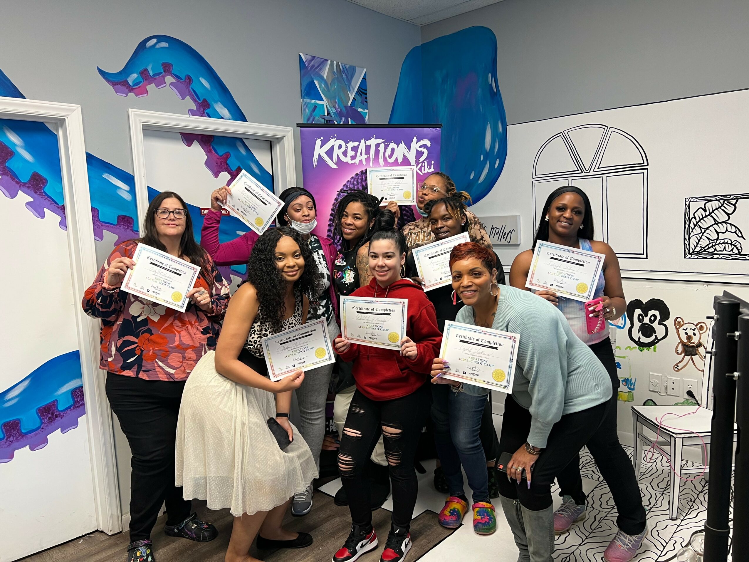 Group of nine people holding certificates in a colorful room with abstract wall art and windows.