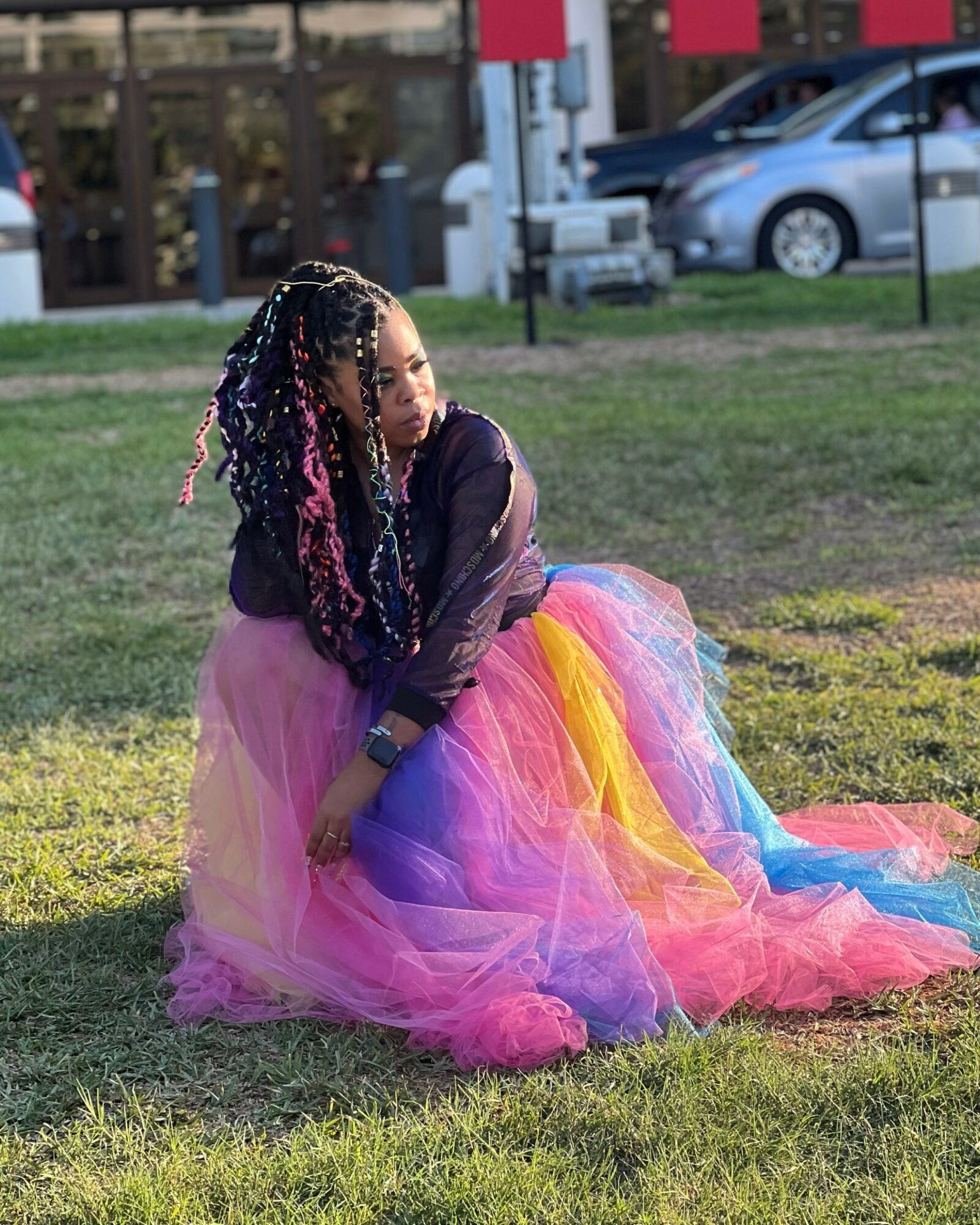 Person sitting on grass wearing a colorful tulle skirt and dark top, with braided hair, outdoors near parked cars.
