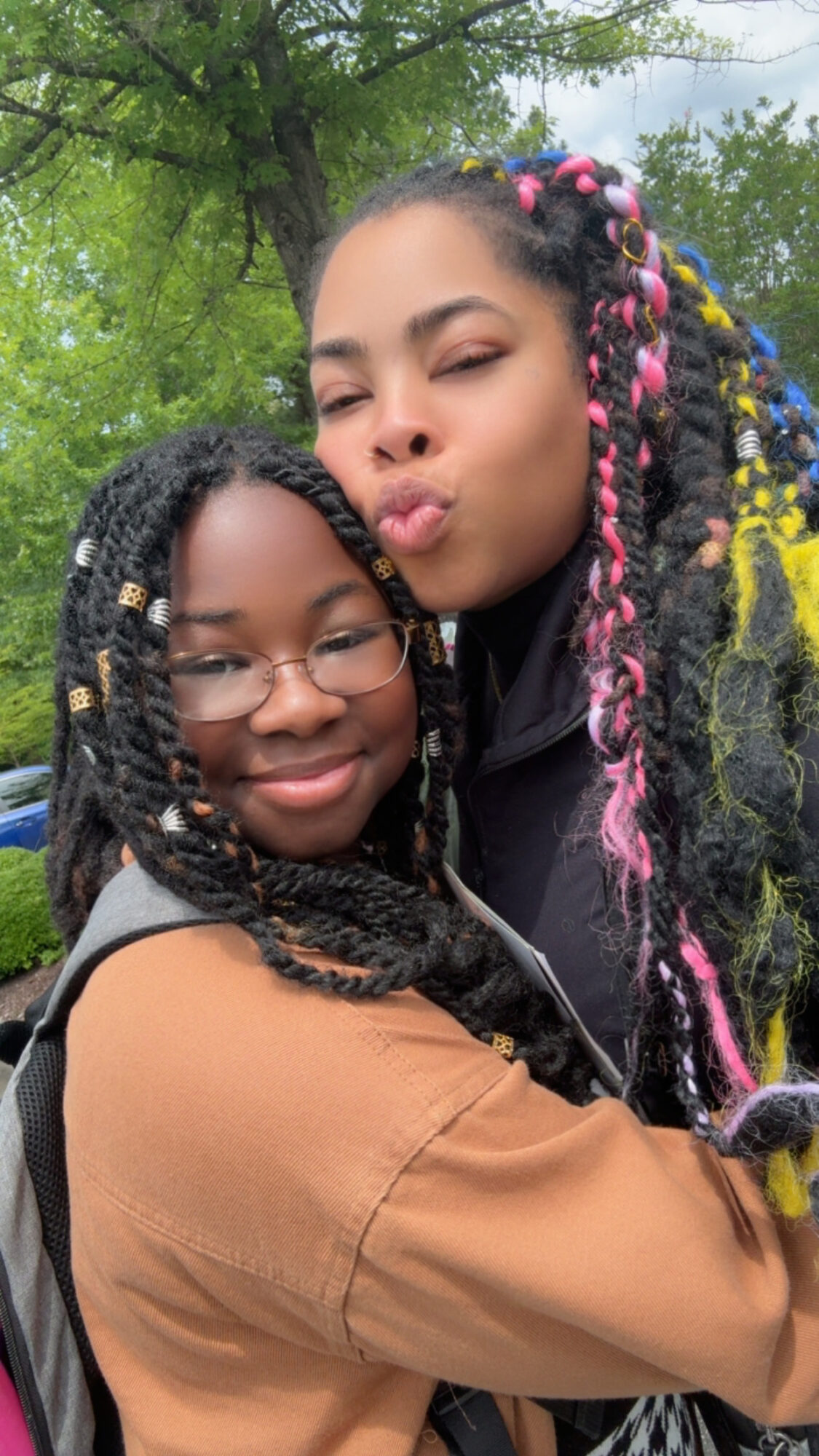 Two young women hugging outdoors, one with glasses and braided hair, the other with colorful braided hair, both smiling.