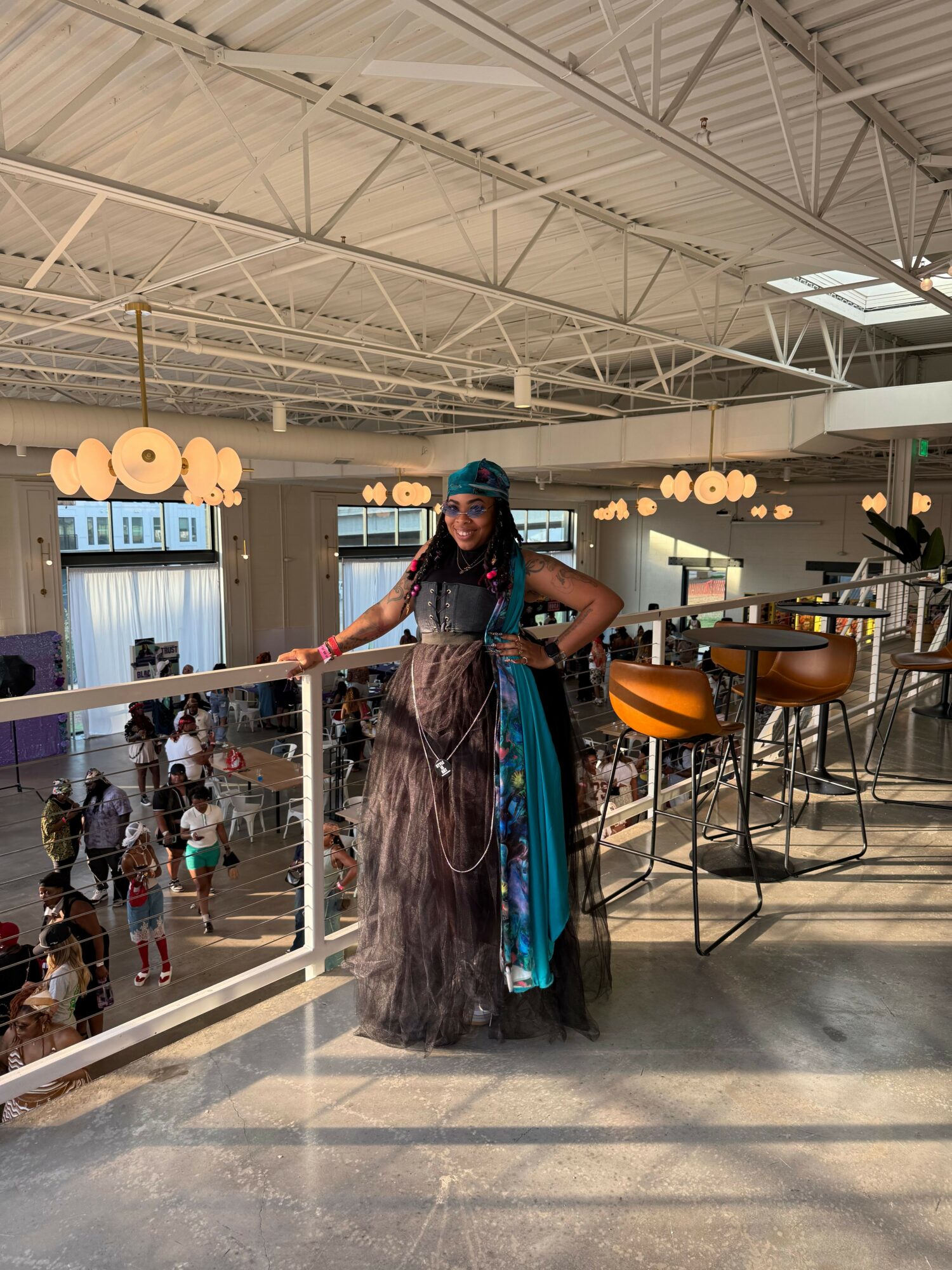 Woman in a long dress and headscarf standing at a table with food, in a spacious indoor setting with tables and chairs.