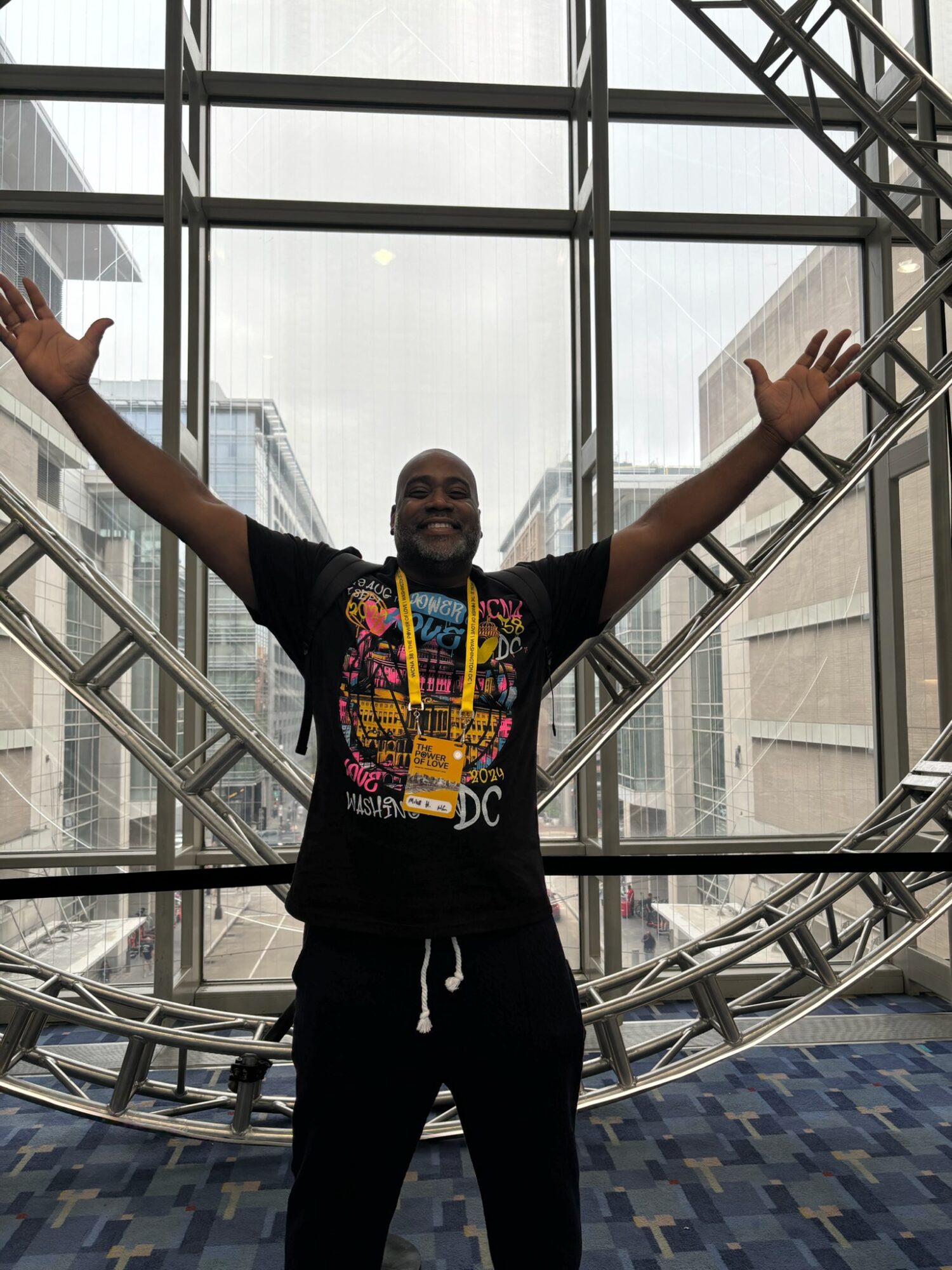 Person with arms raised standing in front of large glass windows and metal framework, smiling, in an indoor setting.
