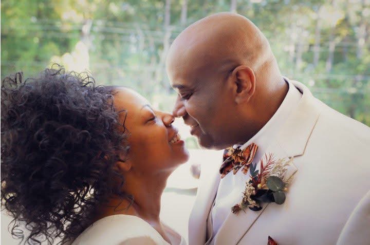 A couple with closed eyes and noses touching, outdoors with trees in the background.