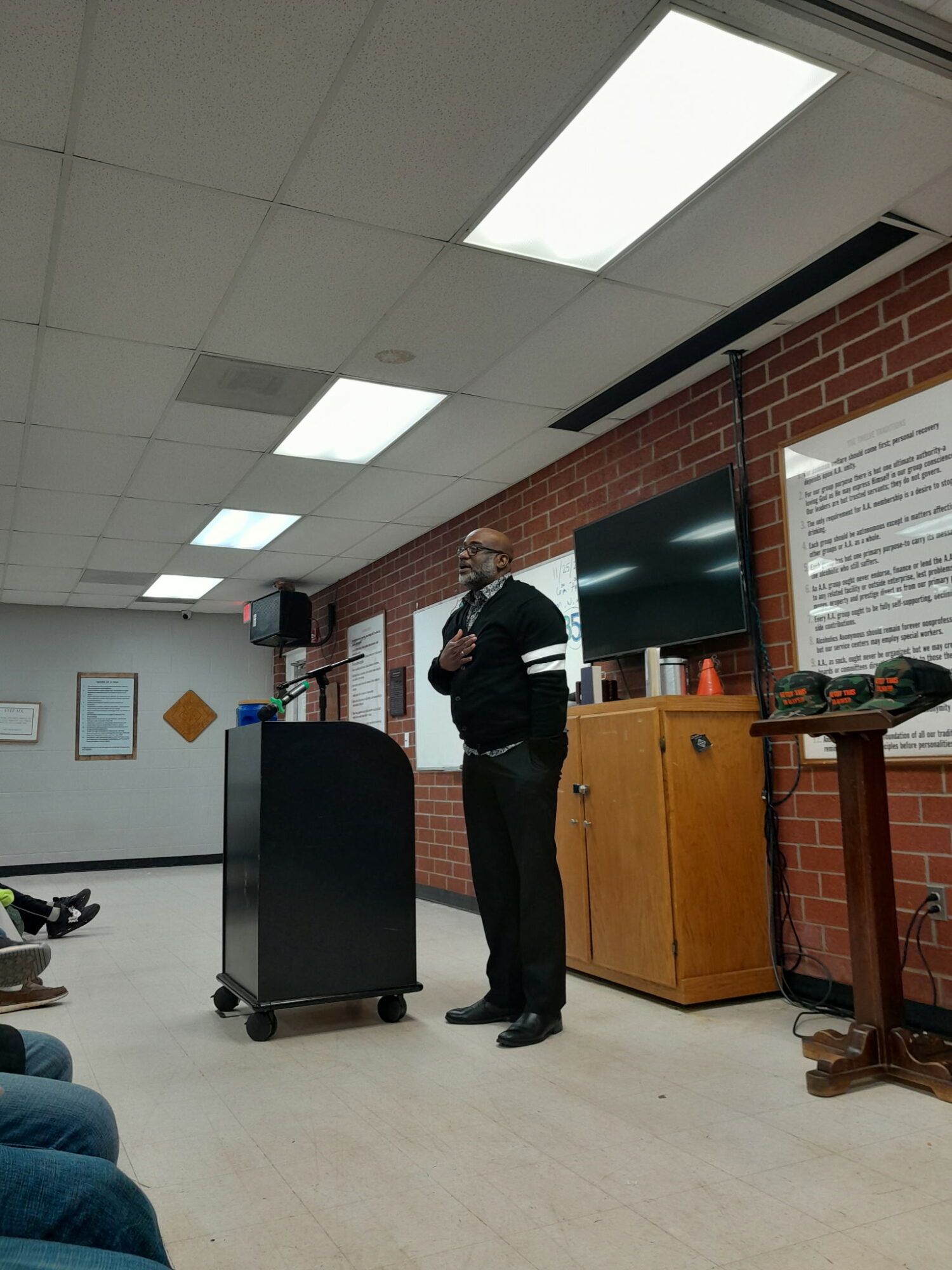 Man standing and speaking in front of a brick wall with a large screen and a wooden cabinet.