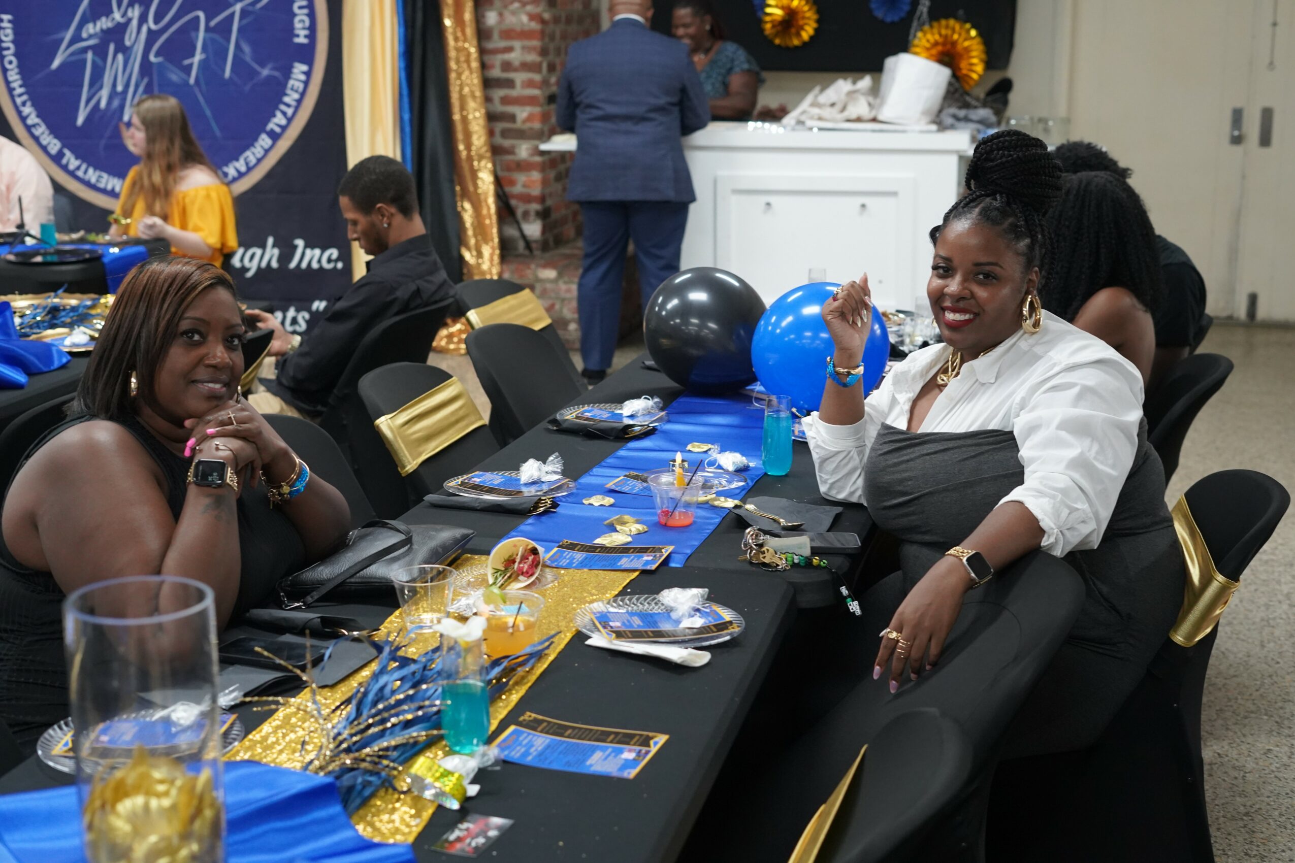 Two women sitting at a decorated table with party favors, balloons, and blue decorations, smiling and waving at the camera.
