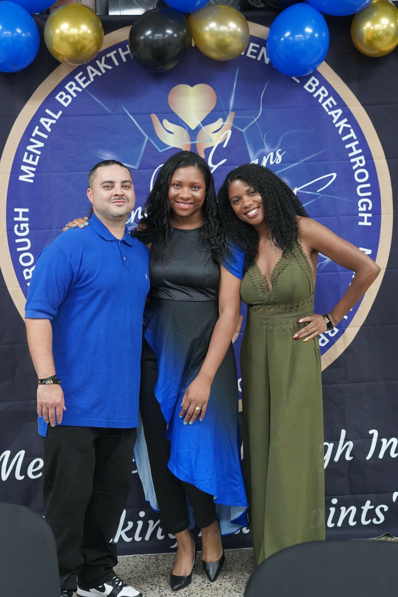 Two women standing in front of a blue circular backdrop with balloons, one in a blue dress and the other in black and green, smiling and posing with a peace sign.