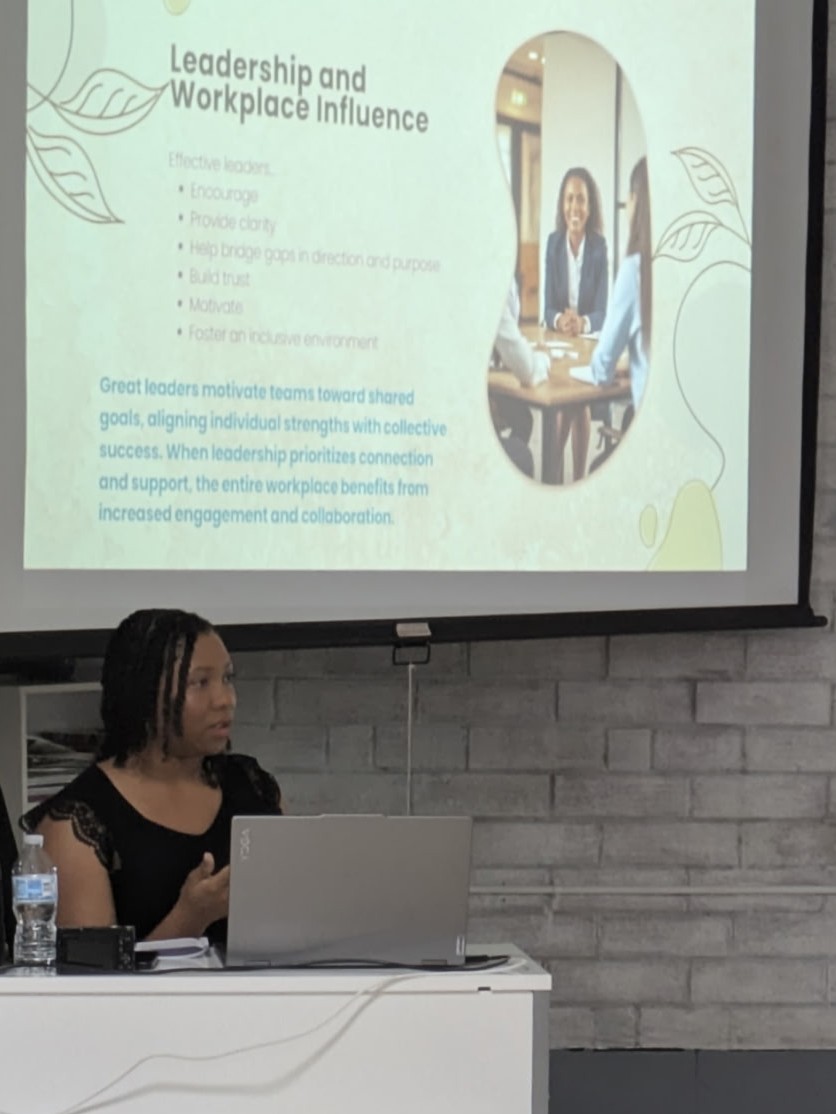 Woman sitting at a desk with a laptop, in front of a presentation slide about leadership and workplace influence, brick wall background.