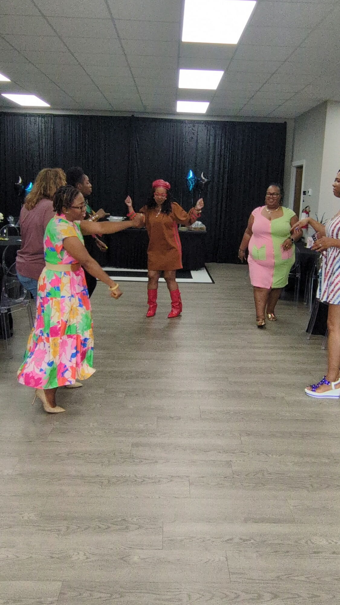 Group of women dancing in a room with a black curtain background, some holding hands, others clapping, wearing colorful dresses.