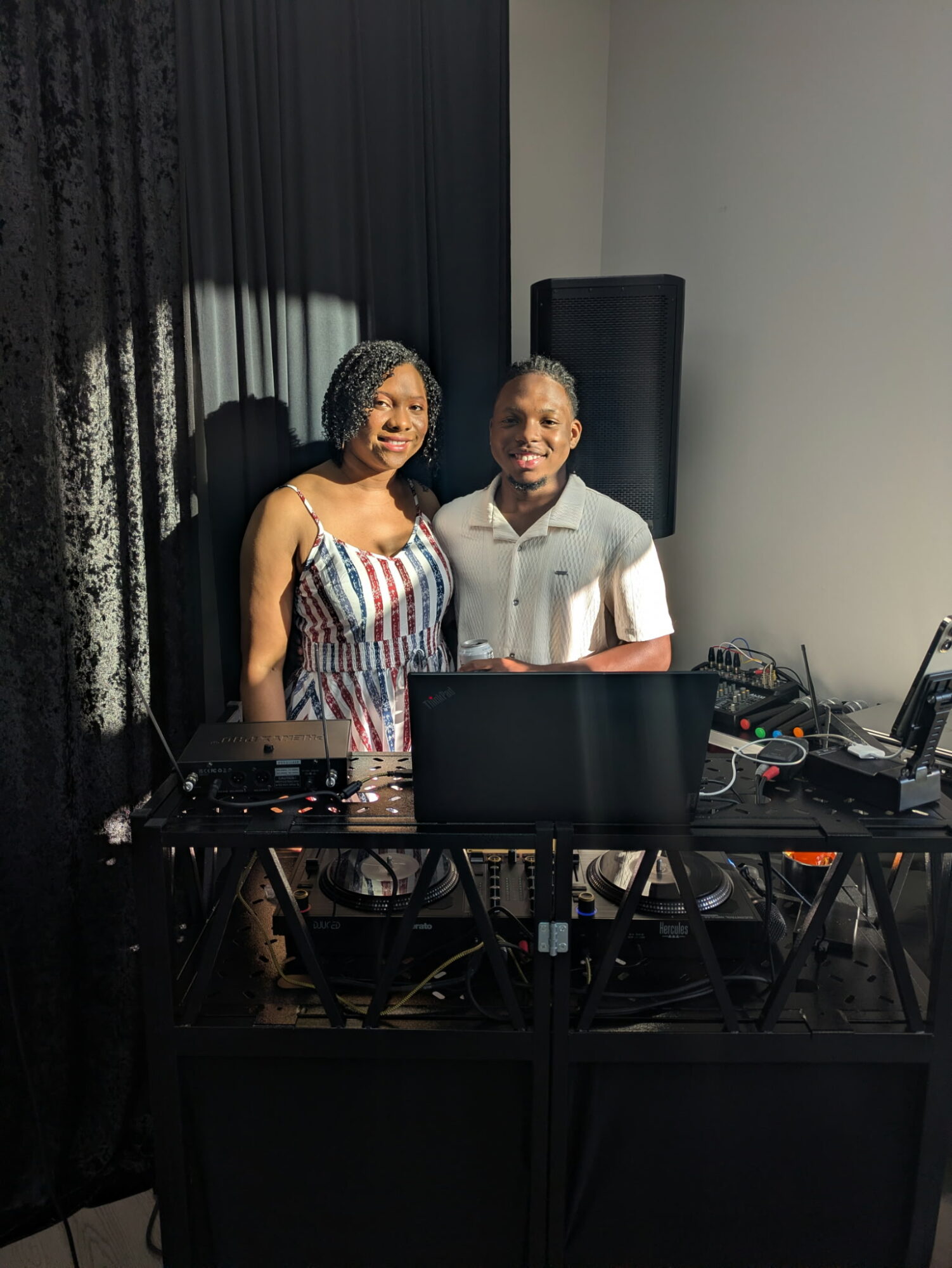Two women stand behind a DJ setup with equipment, black curtains in the background, and a white wall.