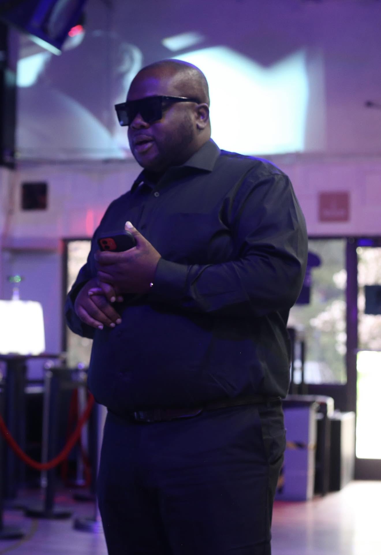 Man wearing sunglasses and black shirt holding a phone indoors, with colorful lighting and blurred background.