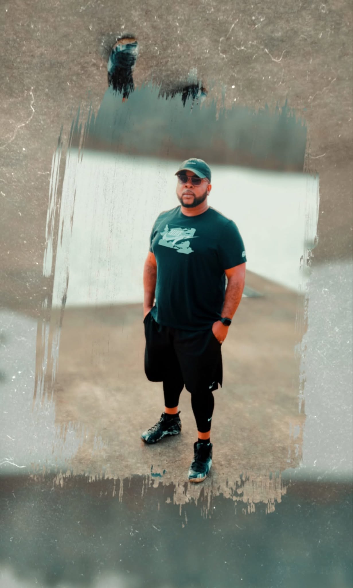 Man wearing a cap, sunglasses, and athletic clothing standing outdoors on a concrete surface.