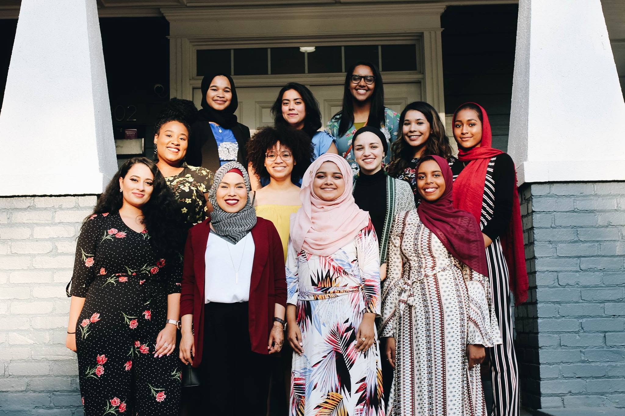 Group of twelve women standing outside a house, smiling, with some wearing hijabs and colorful clothing.