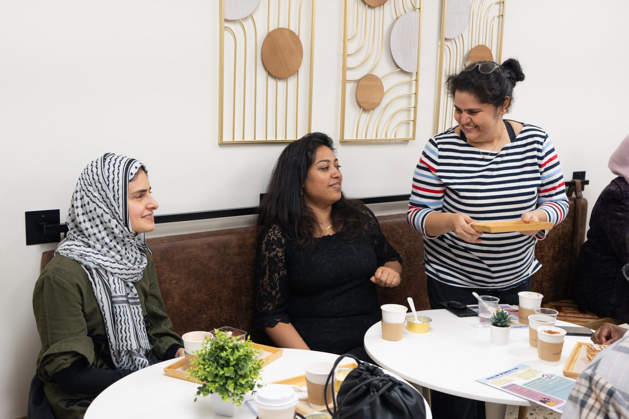Three women sitting at a table, one standing and serving food, in a room with wall art. Bright, casual setting.