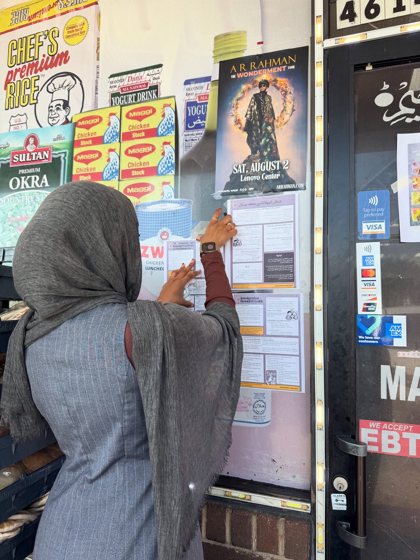Person wearing a gray headscarf and gray coat posting a notice on a bulletin board outside a shop.