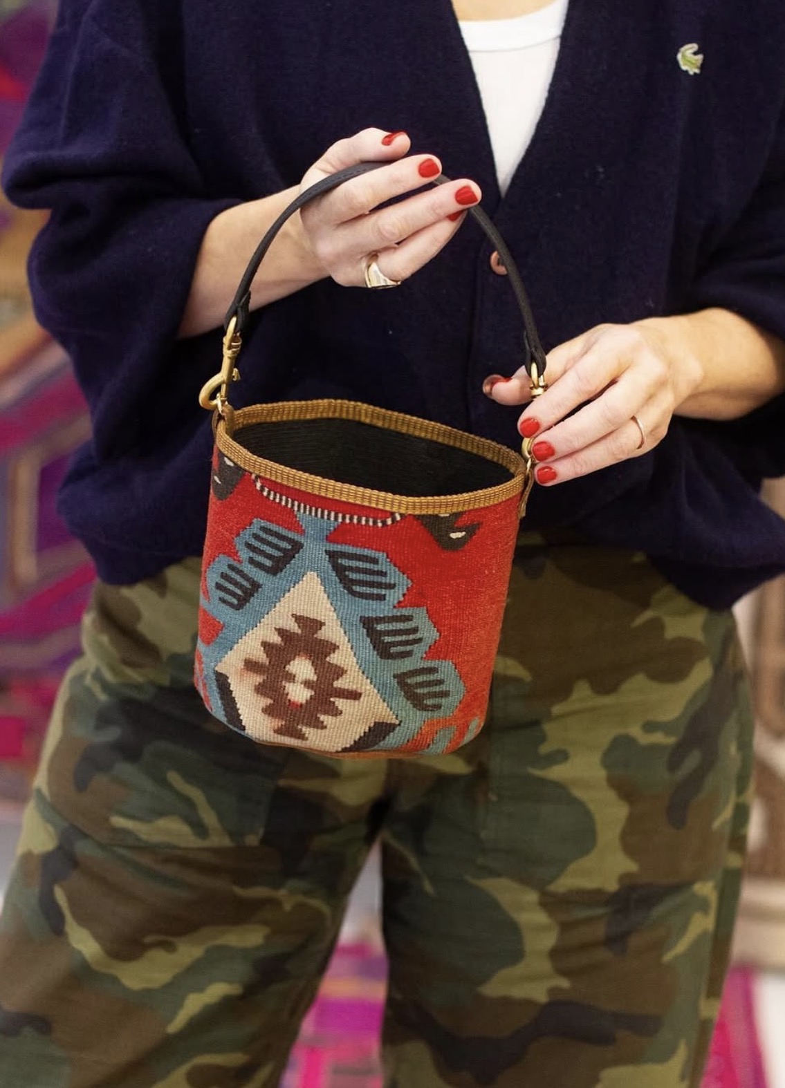 Person holding a colorful patterned bag with a handle, wearing camouflage pants and a dark top, with red nail polish.
