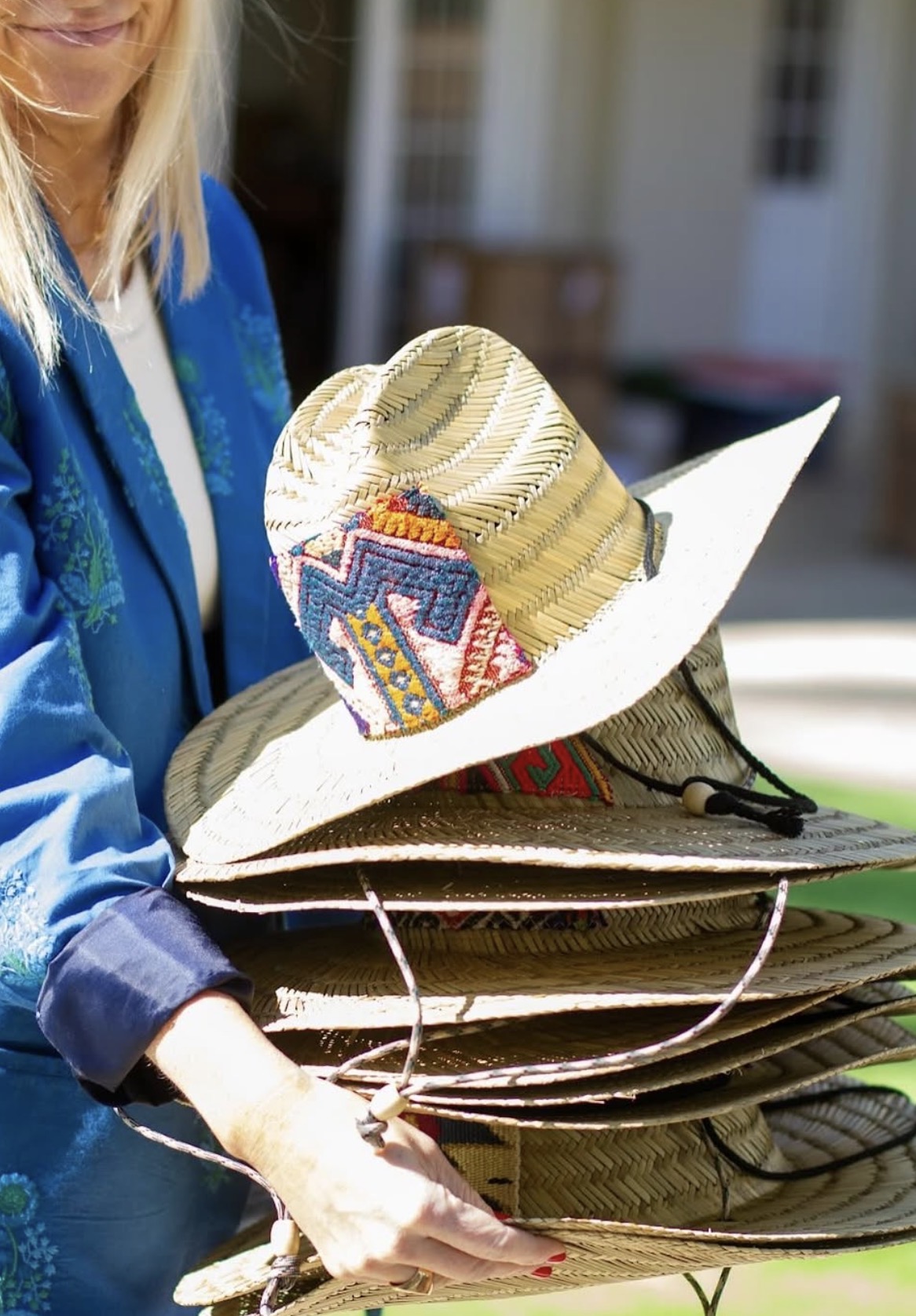 Person holding a stack of straw hats, one with colorful decorations, outdoors with a building in the background.