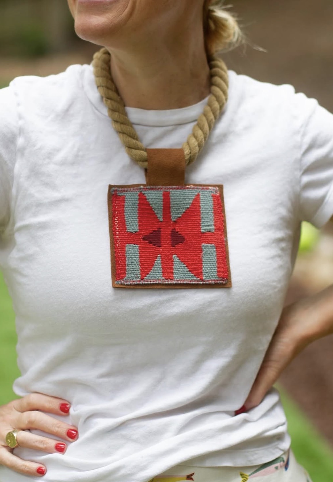 Person wearing a white shirt with a large, colorful square pendant necklace, standing outdoors with one hand on hip.