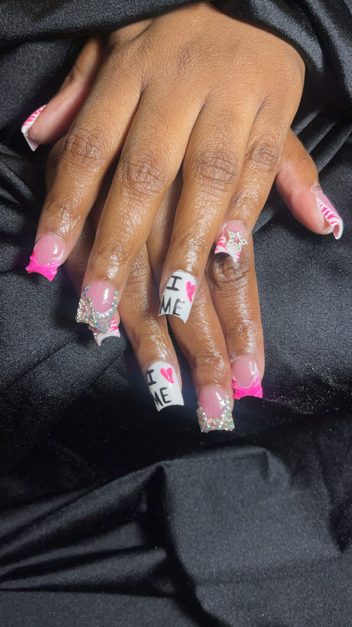 Two hands with decorated nails, one hand resting on the other, nails painted pink with white tips and messages, on black fabric.