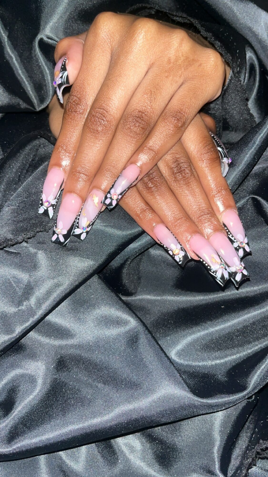 Hands with long, decorated nails resting on black satin fabric, rings on fingers, nails with pink and black designs.