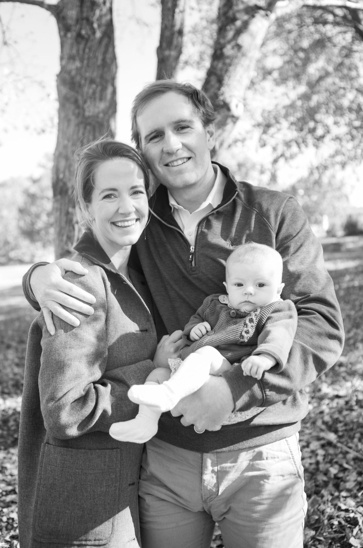 A smiling family of three outdoors, with trees in the background, includes a woman, man, and baby in his arms.