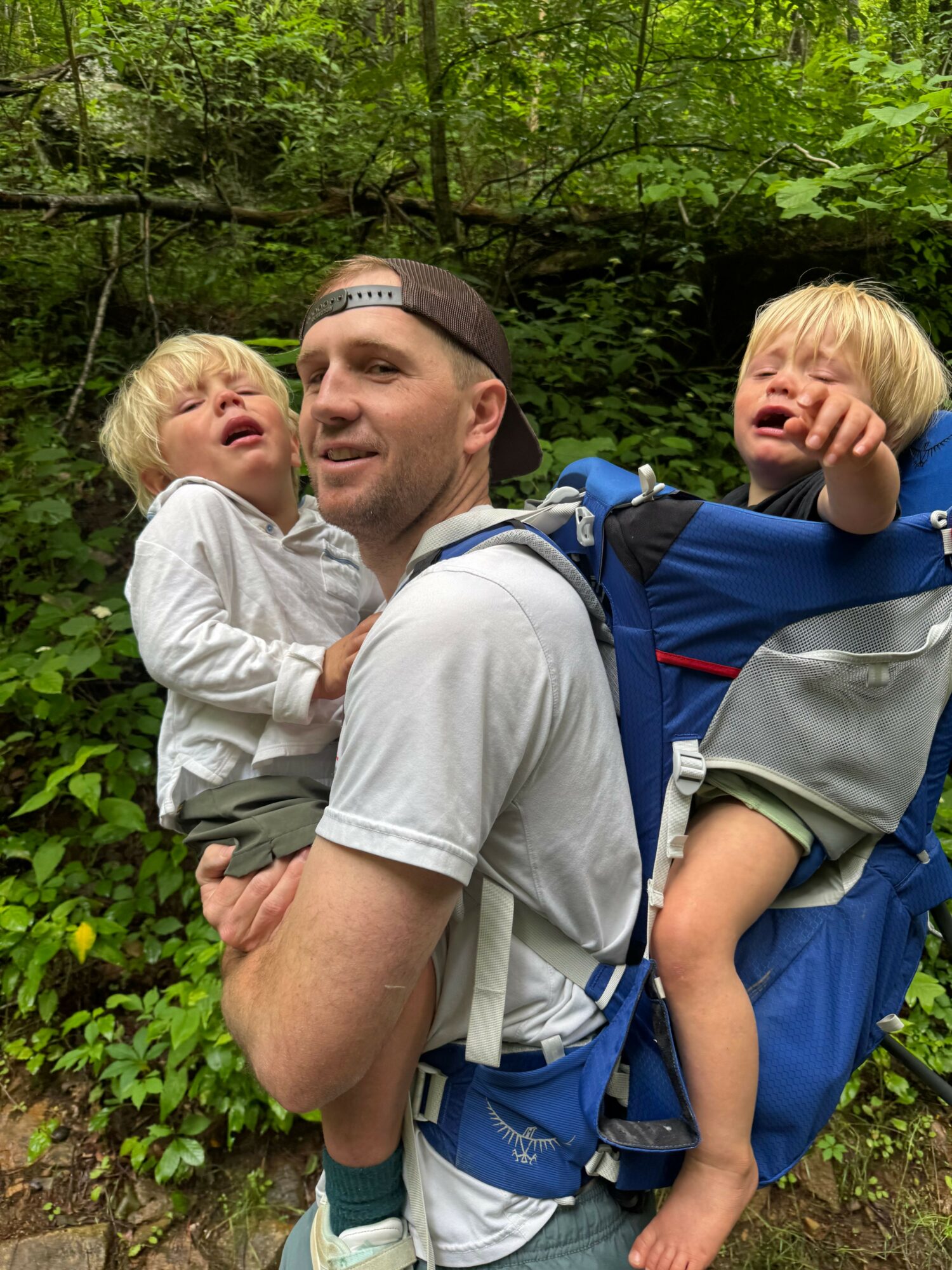 Adult man carrying a child on his back outdoors among green foliage, both smiling and looking at the camera.