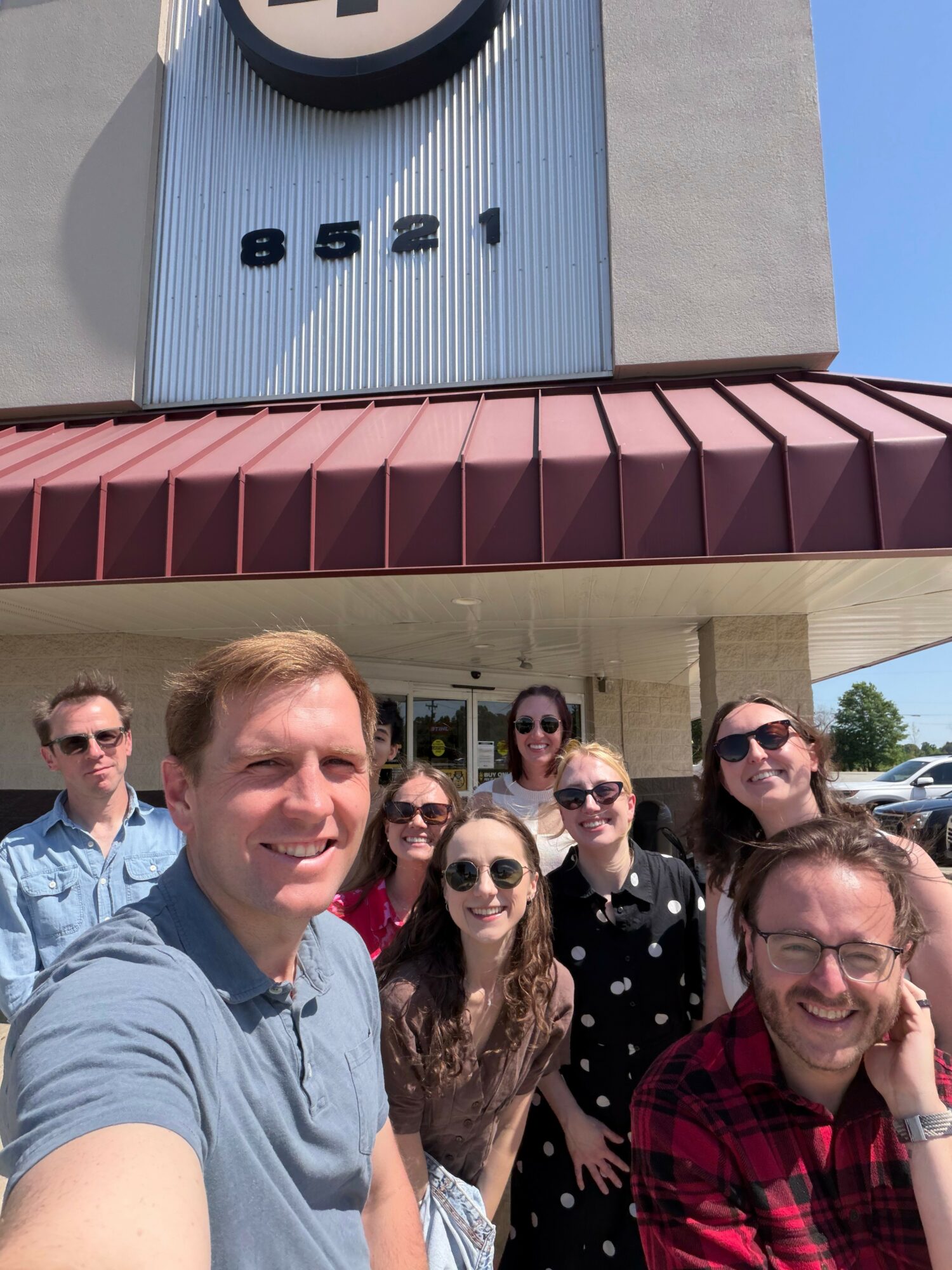 Group of eight people smiling outside a building with a sign displaying the number 8521.