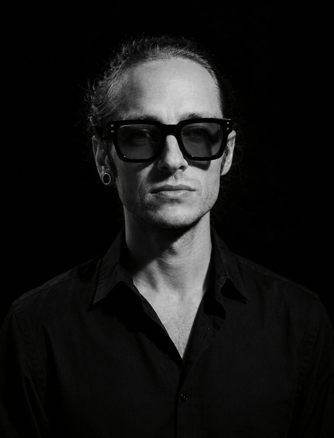 Person with glasses and slicked-back hair wearing a dark shirt against a black background.