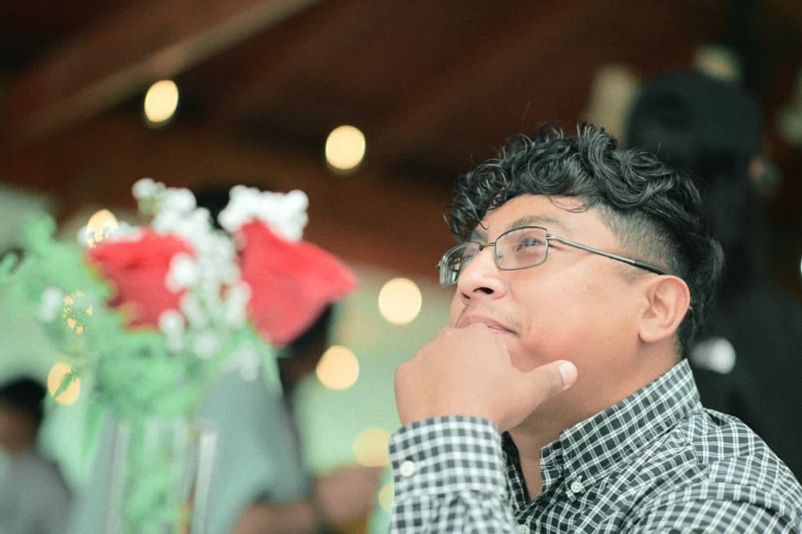Young man with glasses and checkered shirt, hand on chin, in a social setting with blurred background lights.