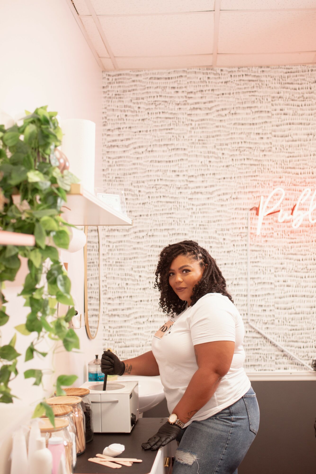 Woman with curly hair wearing a white shirt and jeans, standing in a room with a textured wall and pink neon sign, holding a small object.
