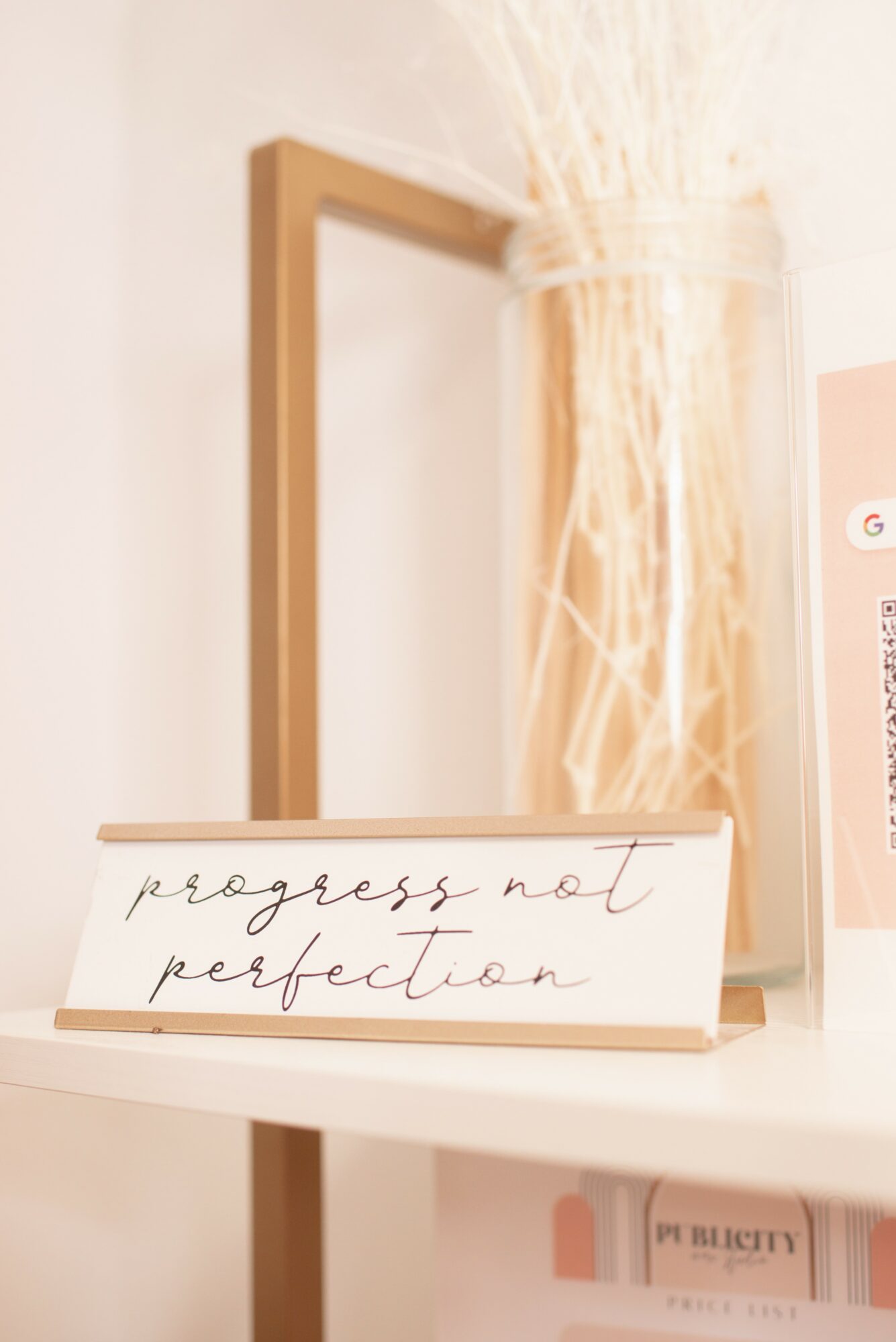 Close-up of a white shelf with a framed mirror, a glass jar with dried plants, and a handwritten note reading 'progress not perfection'.
