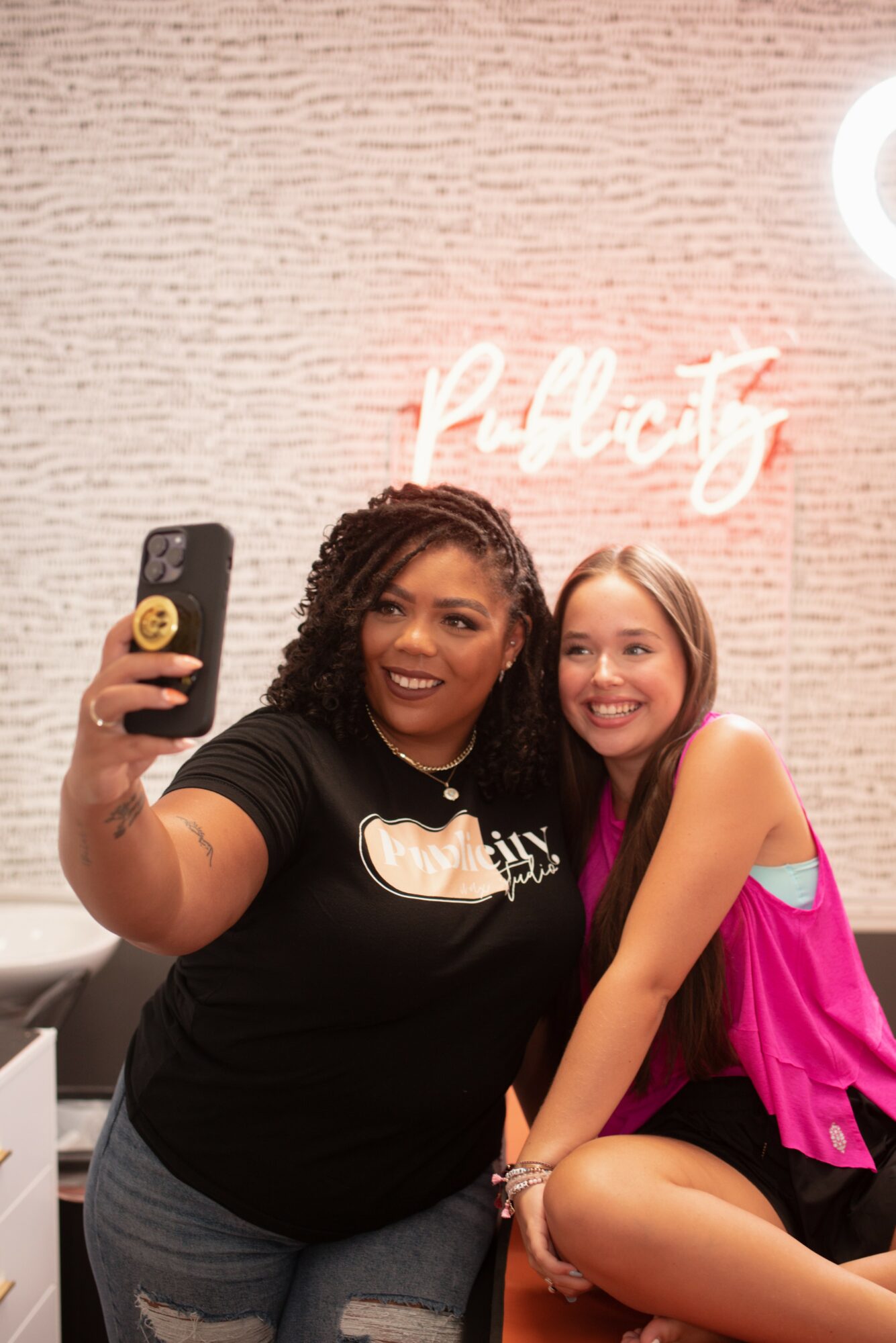 Two women smiling and taking a selfie in front of a wall with a pink neon sign that says 'pretty'.