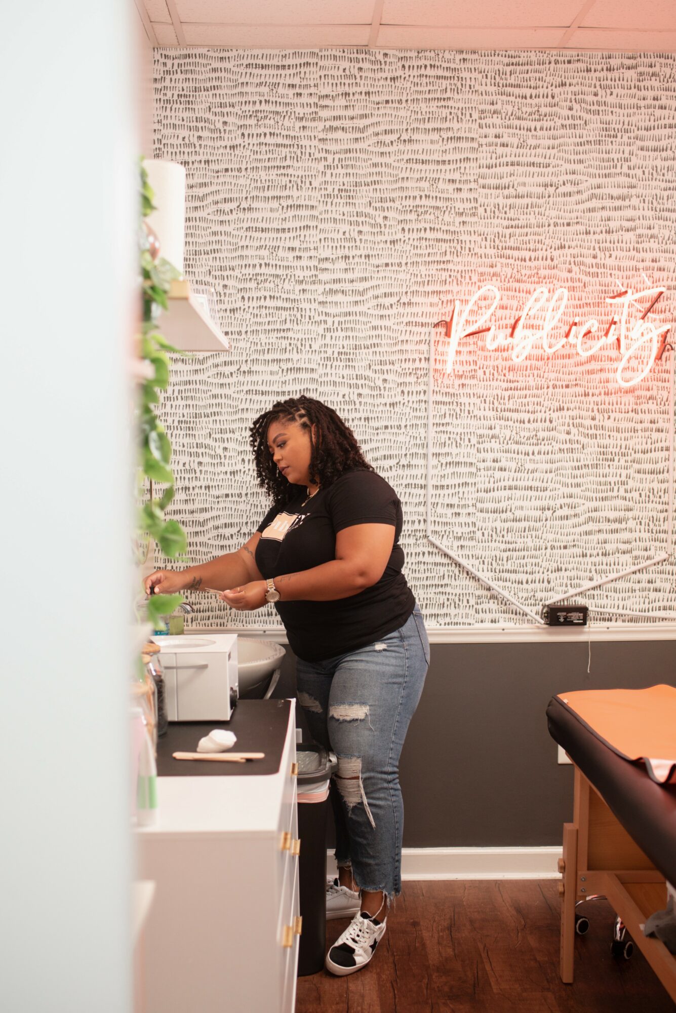 Woman with curly hair in black shirt and jeans standing at a counter, preparing something, in a room with pink and gray walls.
