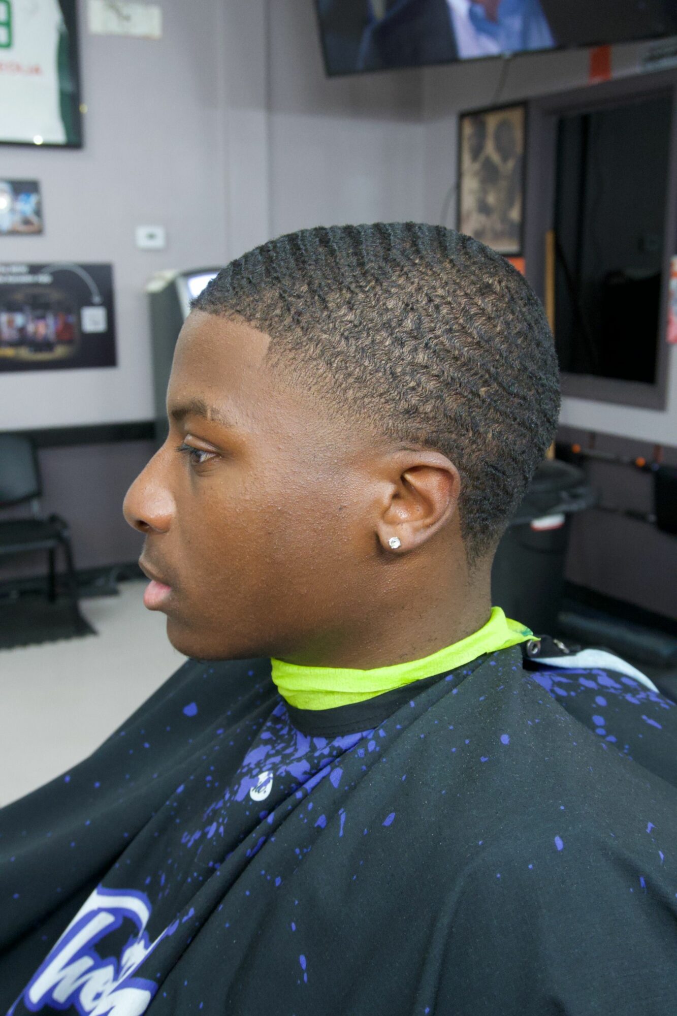Side view of a young person with a short, textured haircut, wearing a black shirt with blue and green accents, indoors.