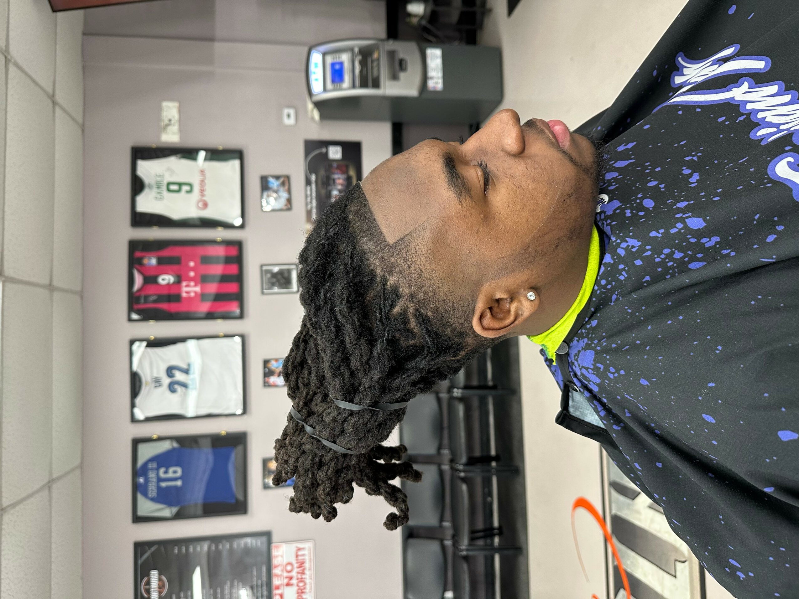 Side view of a man with styled dreadlocks in a barbershop chair, with framed sports jerseys on the wall behind him.